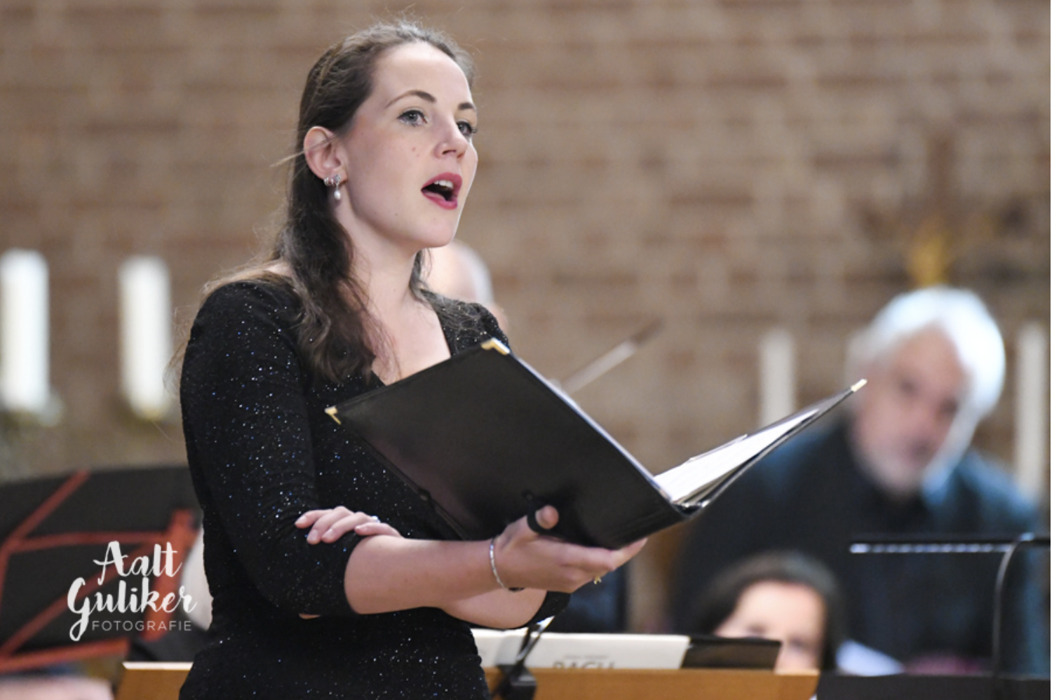 Een vrouw zingt tijdens een zanguitvoering, met een muziekblad in haar hand. Achter haar bevinden zich enkele kaarsen en een muzikale opgebouwd. Op de achtergrond is een man te zien die op een piano speelt.