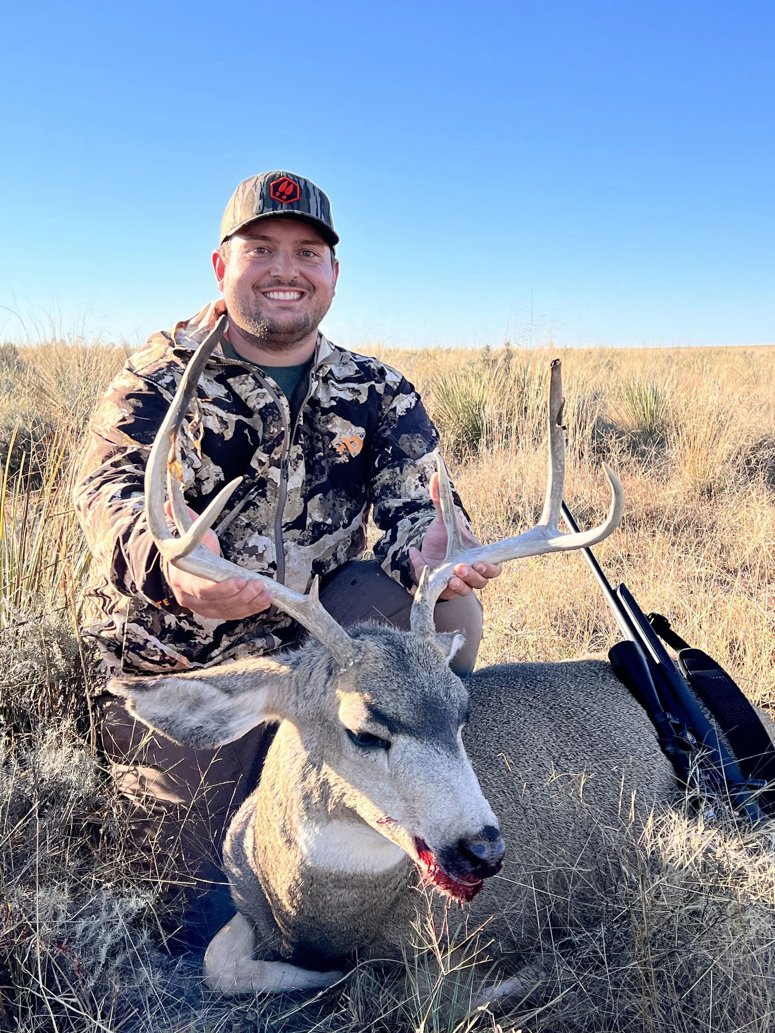 A man in camouflage clothing and a baseball cap kneels in a field of tall grass, holding a large deer with antlers. The deer appears to be recently hunted, with blood visible on its nose. The man is smiling and holding the deer's antlers, with a rifle resting on the ground beside him. The sky is clear and blue.