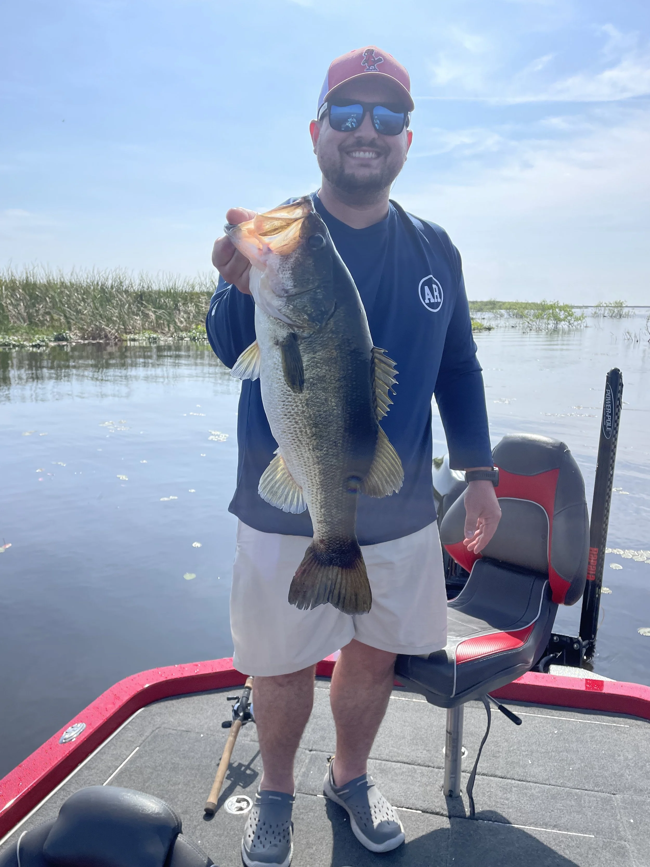 A man standing on a boat holding a large fish, smiling, wearing sunglasses, a red hat, a navy shirt with a circular logo, white shorts, and gray Crocs, with water, reeds, and a blue sky in the background.