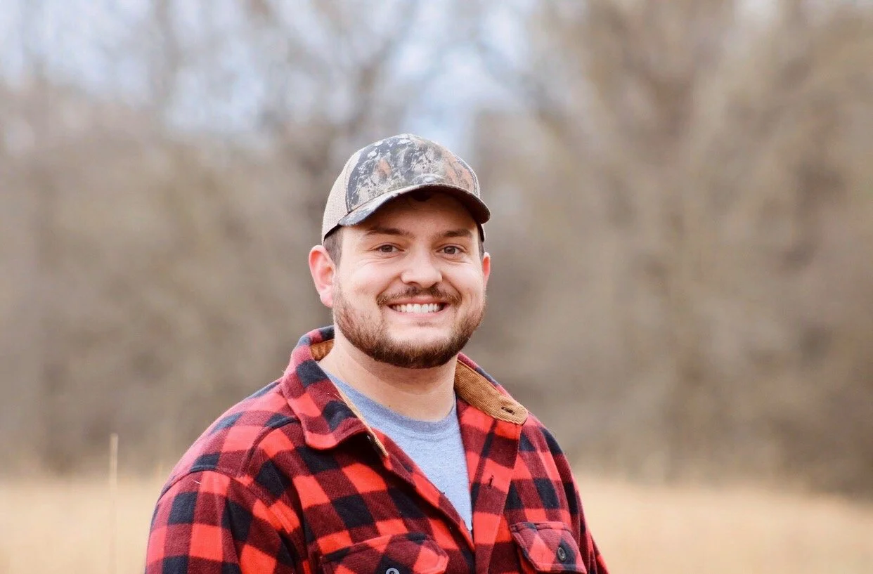 A smiling man in a red and black plaid shirt and a camouflage cap stands outdoors, with a blurred natural background of trees and grass.