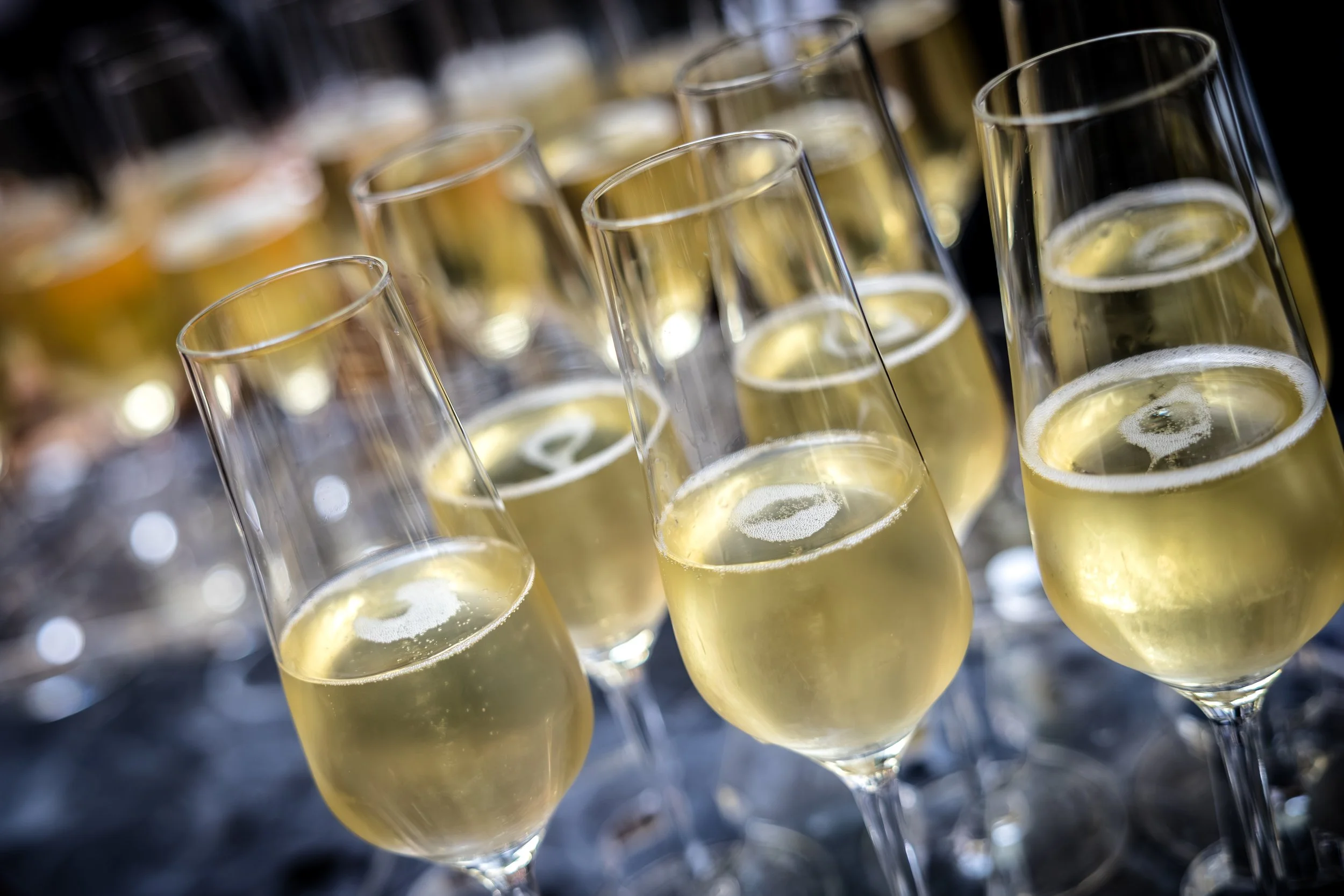 Multiple champagne glasses filled with sparkling wine, tilted slightly and arranged in rows on a dark surface.
