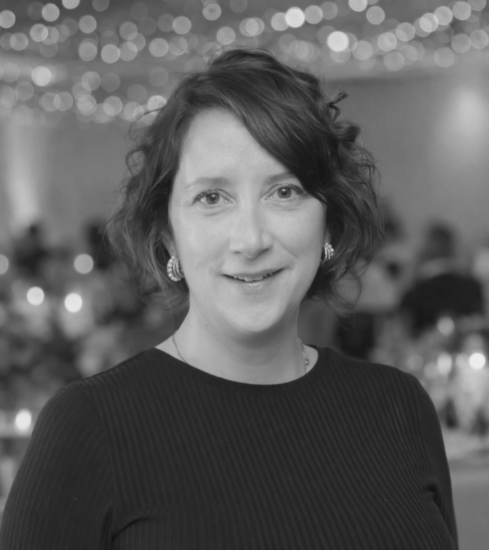 A woman with short wavy hair and earrings, smiling at the camera, in a setting decorated with string lights and blurred background indicating a festive or formal event.