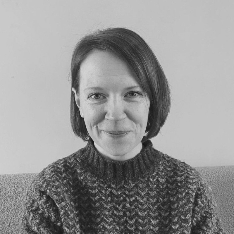 Black and white photo of a smiling woman with short hair, wearing a knitted sweater, sitting in front of a plain wall.