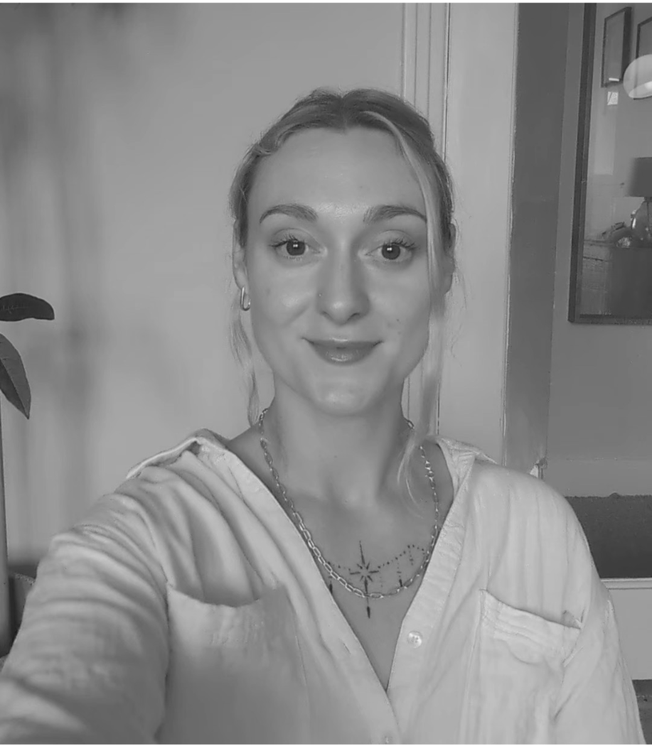 A woman with light-colored hair taking a selfie indoors, smiling at the camera. She is wearing a light-colored shirt and a necklace.