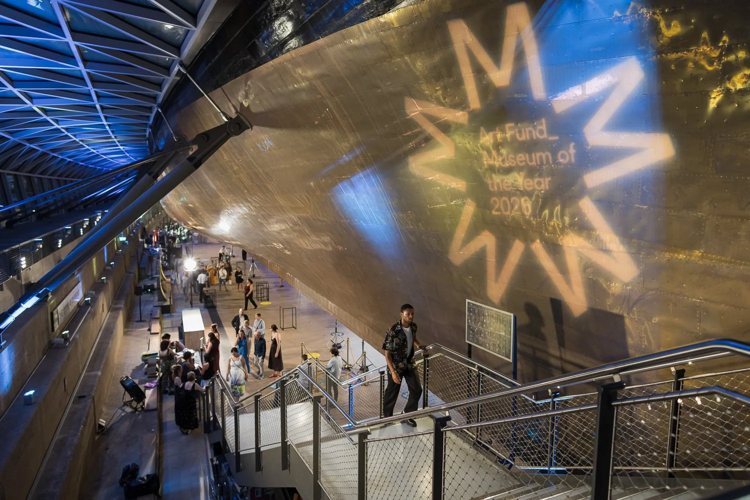 Interior of the Museum of the Future in Dubai, showing visitors ascending a staircase, with a large reflective golden structure and a projection of a star-shaped logo on the wall.