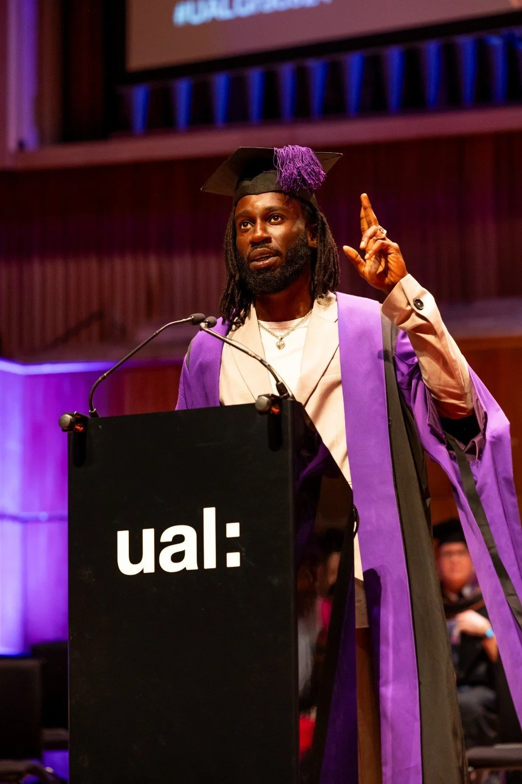 A man speaking at a podium during a graduation ceremony. He is wearing graduation robes and a cap with purple tassel. The background features a large screen and wood paneling.