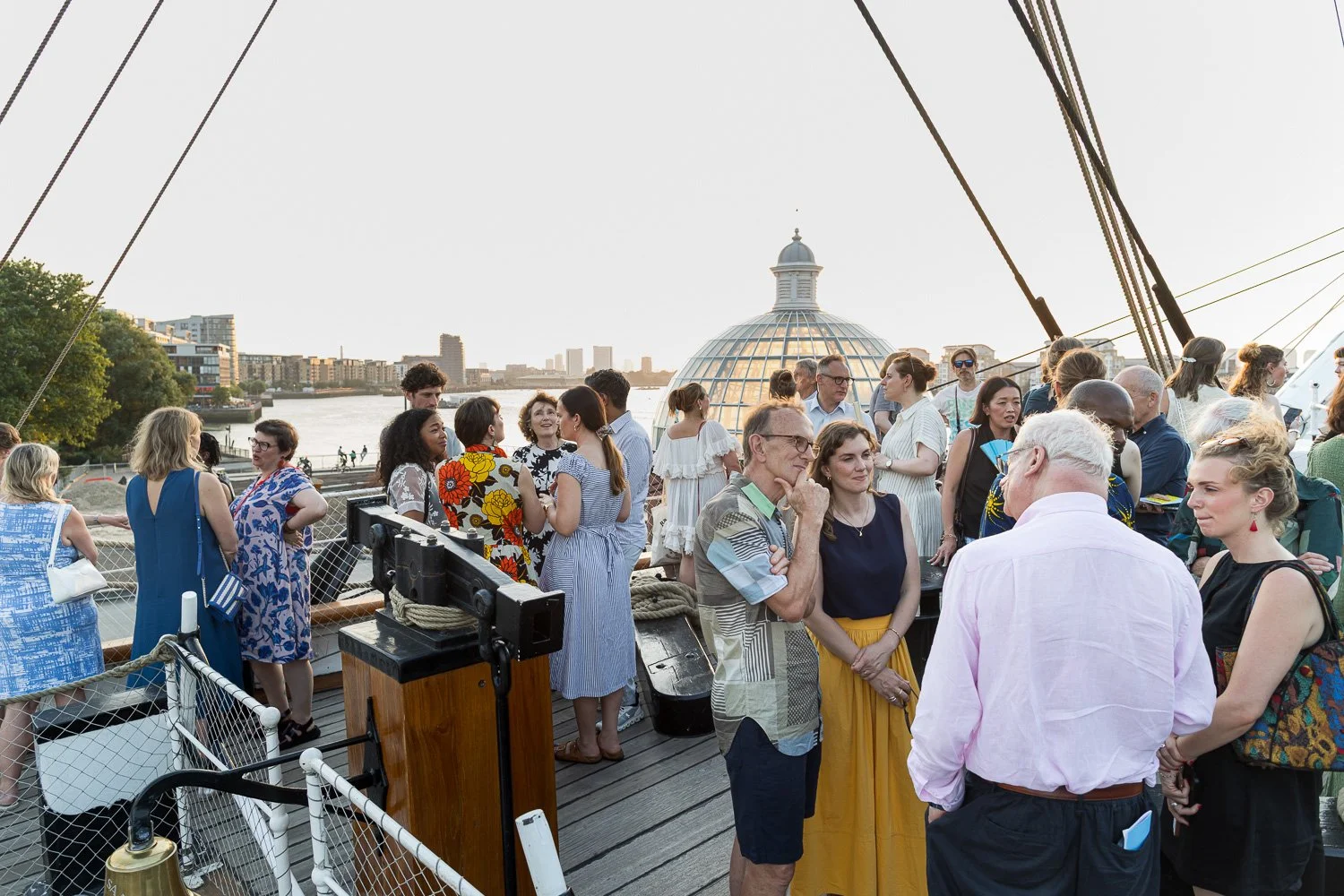 A group of people gathered on a ship's deck with a city skyline and river in the background, during sunset.
