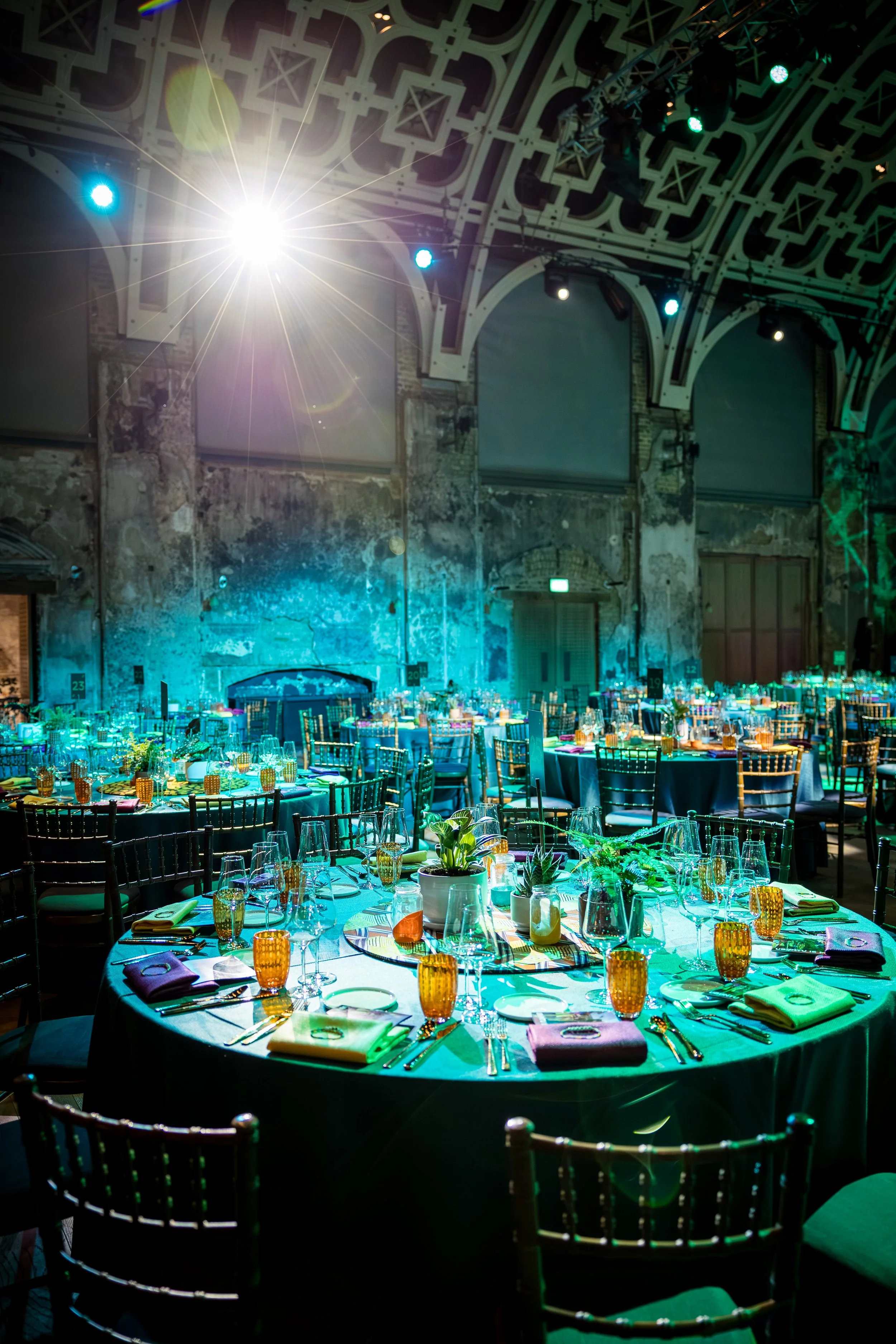 A decorated banquet hall with round tables set with glassware, napkins, and centerpieces, illuminated by colorful ambient lighting and a bright spotlight on the ceiling.
