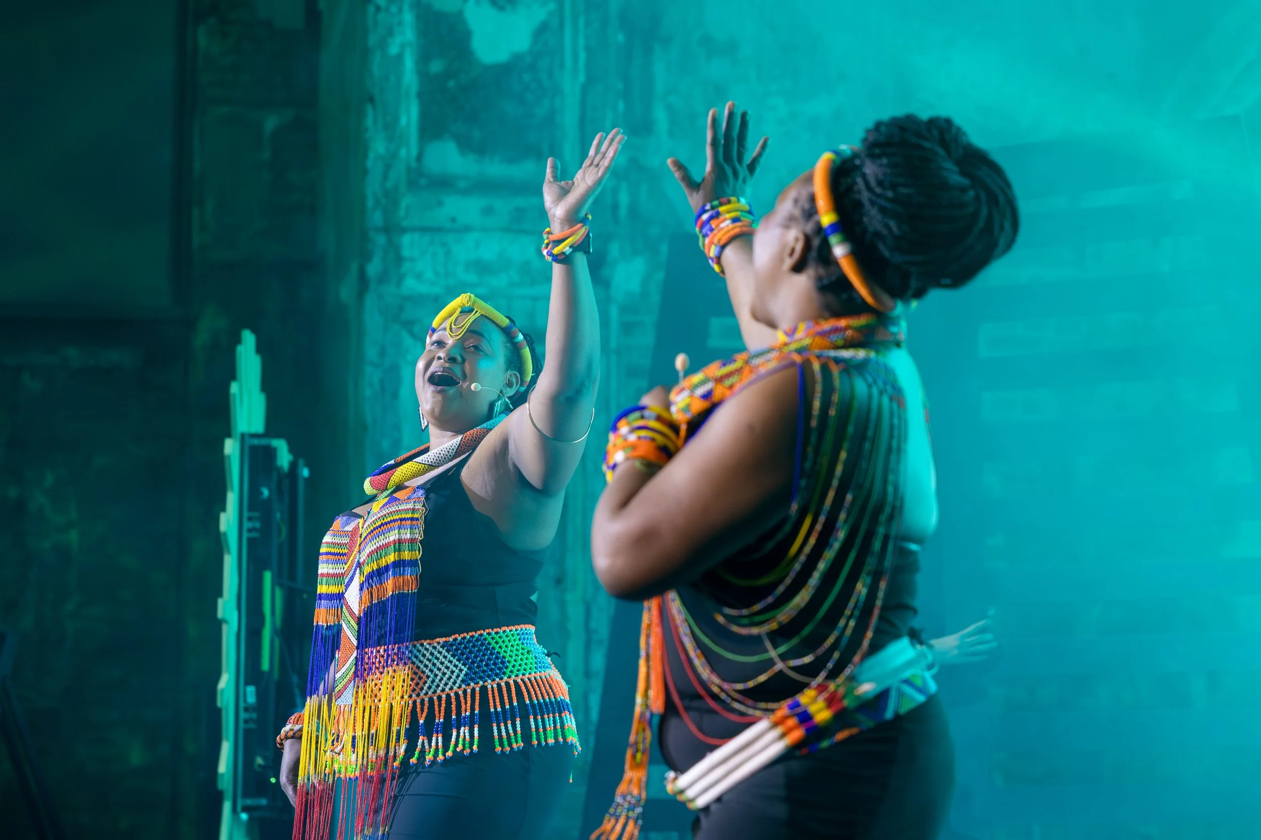 Two women wearing traditional colorful African attire are performing a dance or gesture on stage, with one woman reaching up with her hand while the other watches. The background is dark with blue lighting.