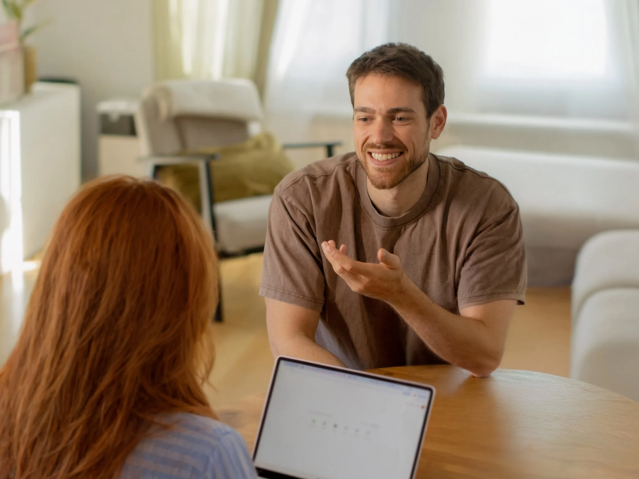 Smiling man gestures while speaking with a red-haired woman across a table with an open laptop.