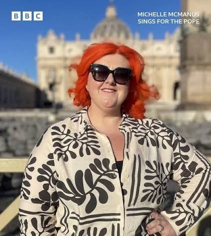 A woman with bright red hair and large sunglasses standing outdoors in front of a historic building, smiling. She is wearing a patterned black and white jacket. The image includes BBC branding and the text 'Michelle McManus Sings for the Pope'.