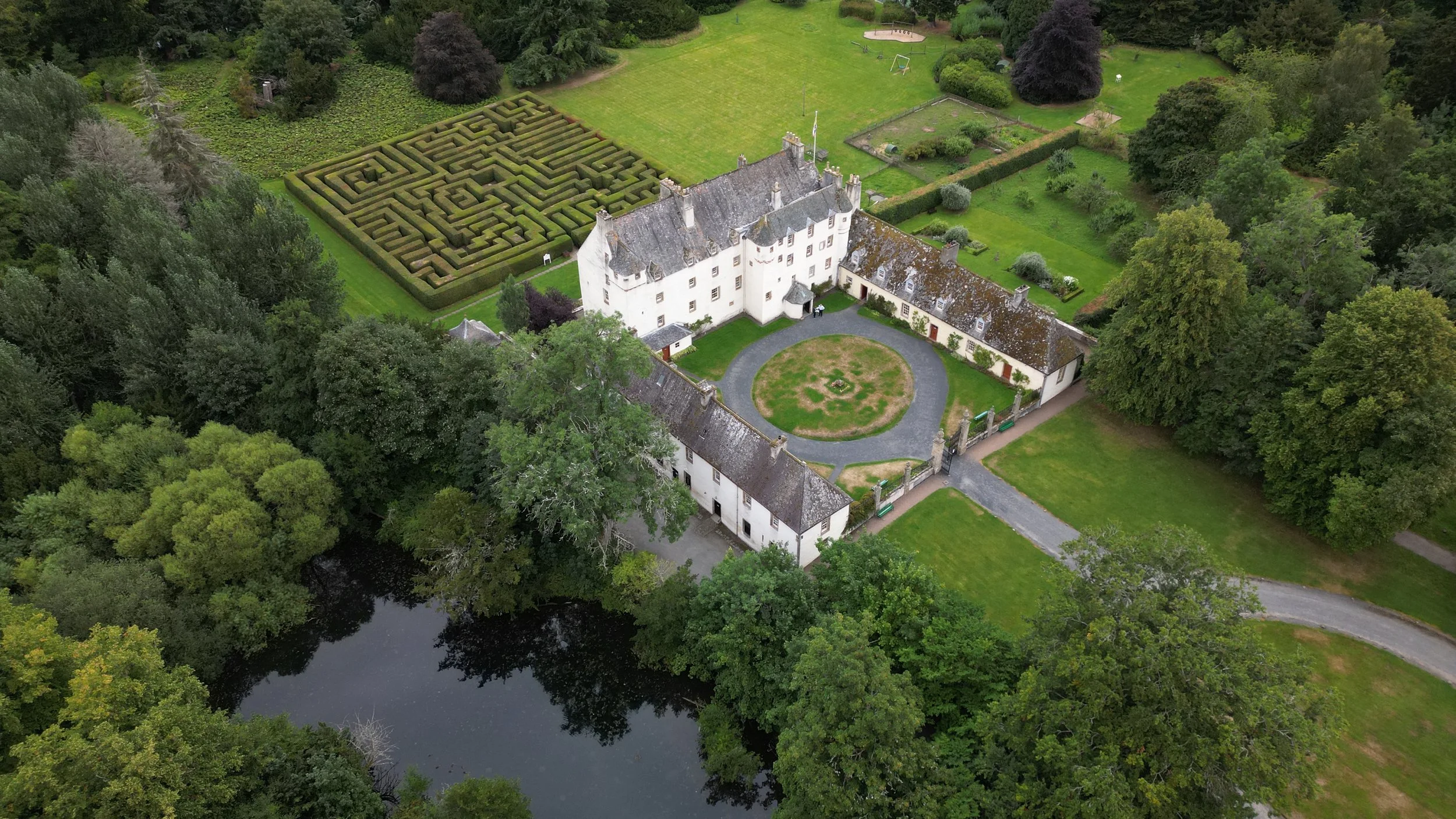 Aerial view of a large white castle-style house with a circular driveway, surrounded by lush green trees, a maze garden, and a river.