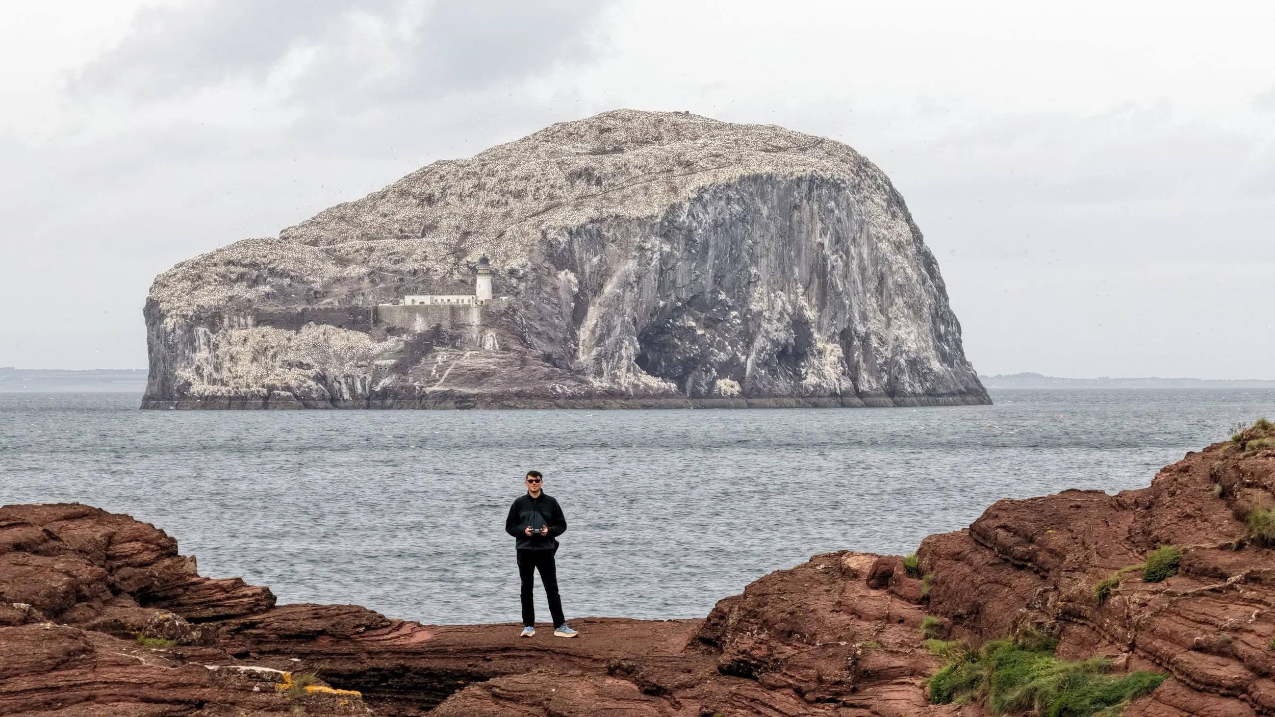 A person standing on rocky shoreline in front of the ocean with large island featuring a lighthouse on a cliff in the background.