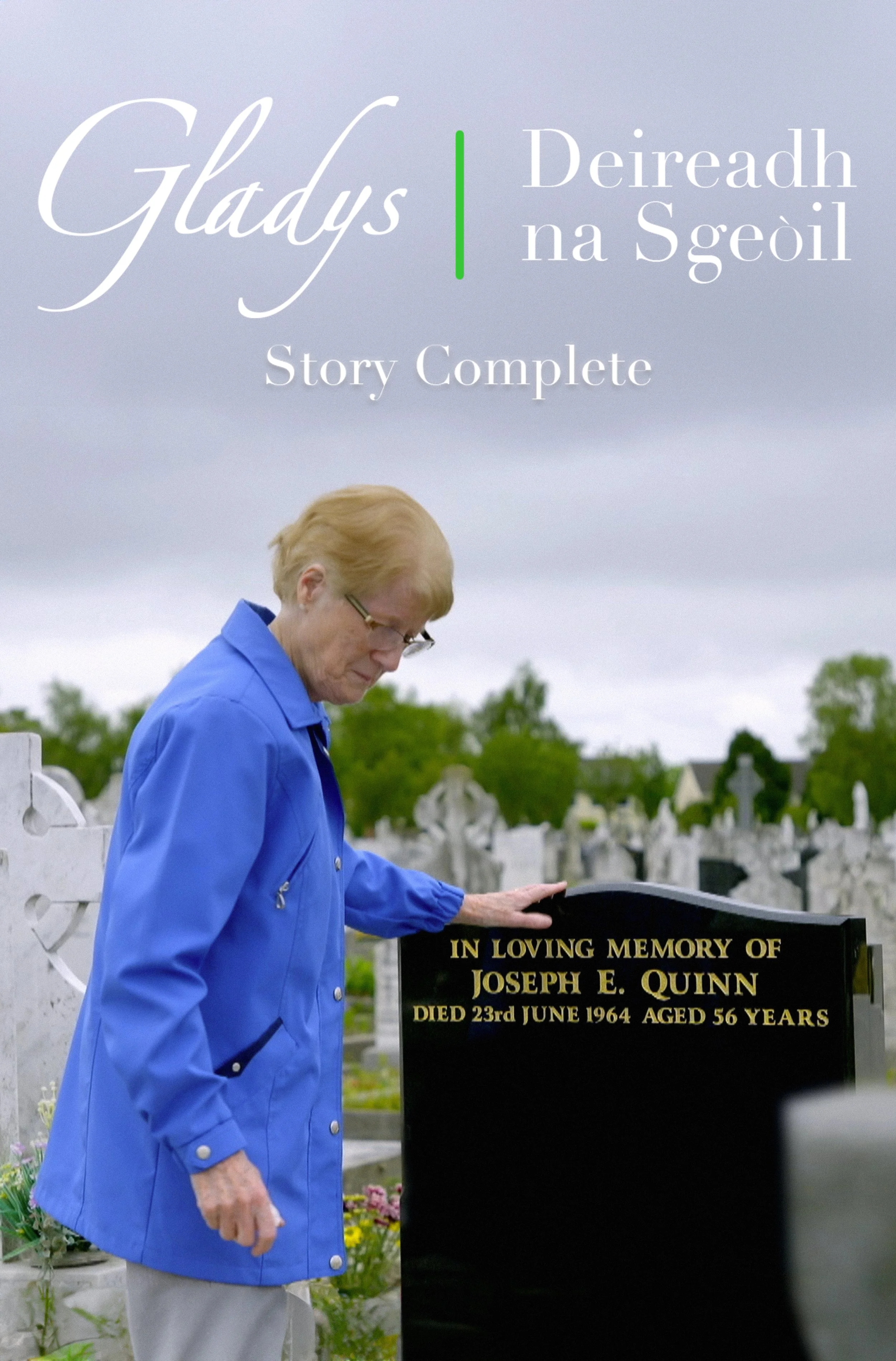 An elderly woman with short blonde hair and glasses wearing a blue jacket, standing in a graveyard, placing her hand on a black memorial gravestone that reads 'In Loving Memory of Joseph E. Quinn, Died 23rd June 1964, Aged 56 Years'.