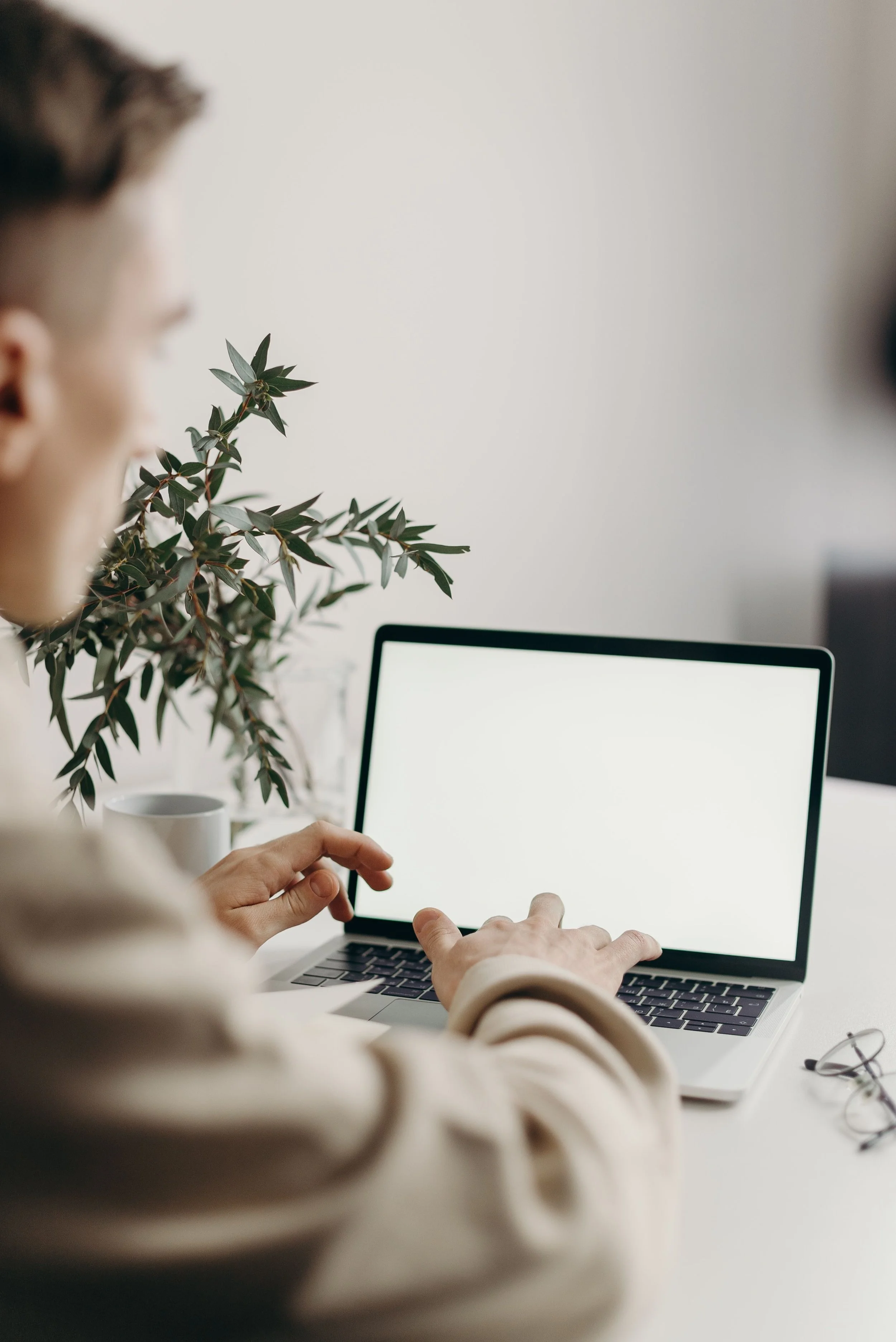 Person in a beige sweater working on a laptop with a blank screen, sitting at a white desk with a potted plant and reading glasses.