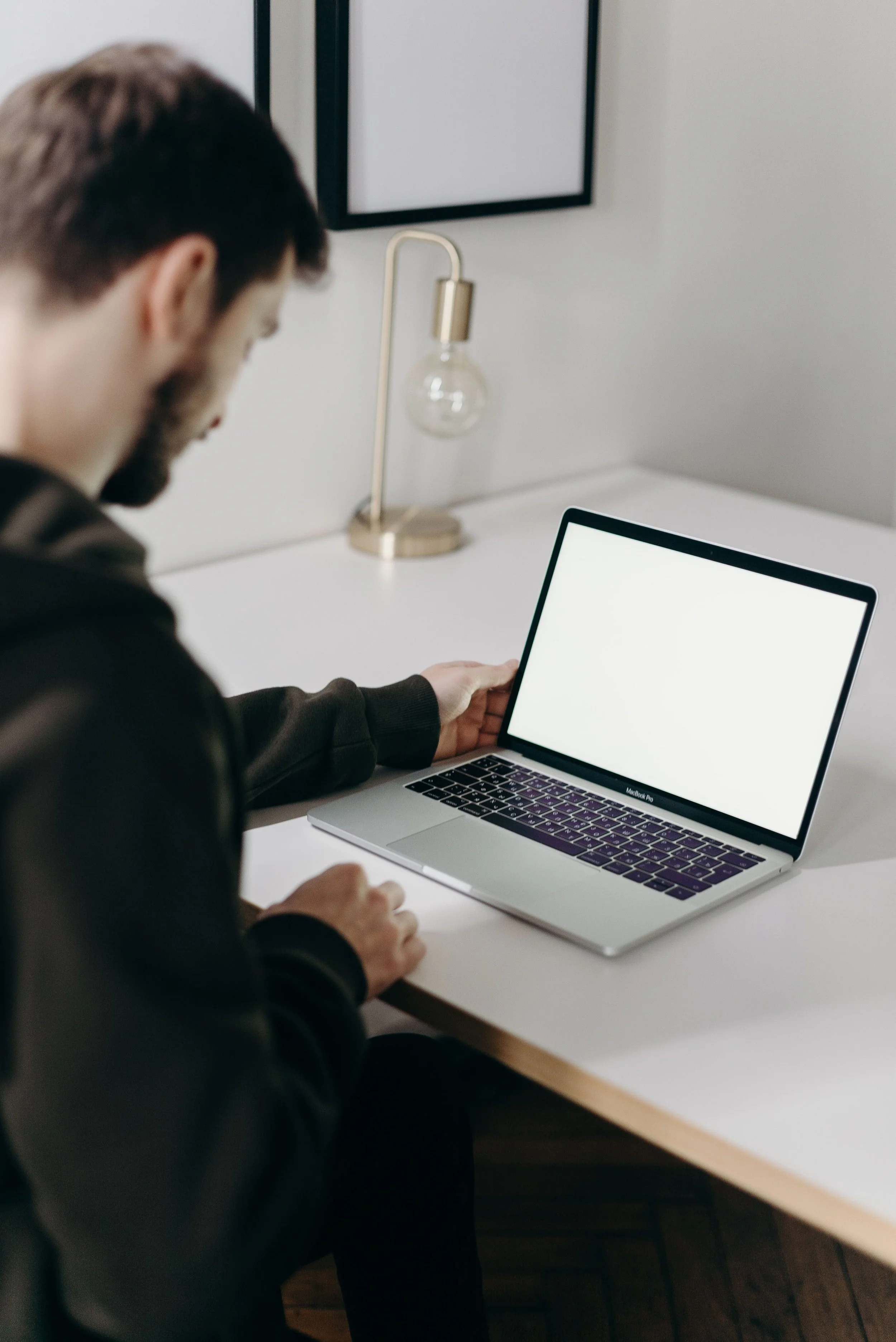 A man with dark hair and a beard sitting at a desk with a silver MacBook Pro laptop. He is wearing a black hoodie and appears to be working or looking at the laptop screen. There is a white wall with framed artwork and a modern desk lamp with a gold base and a clear bulb in the background.
