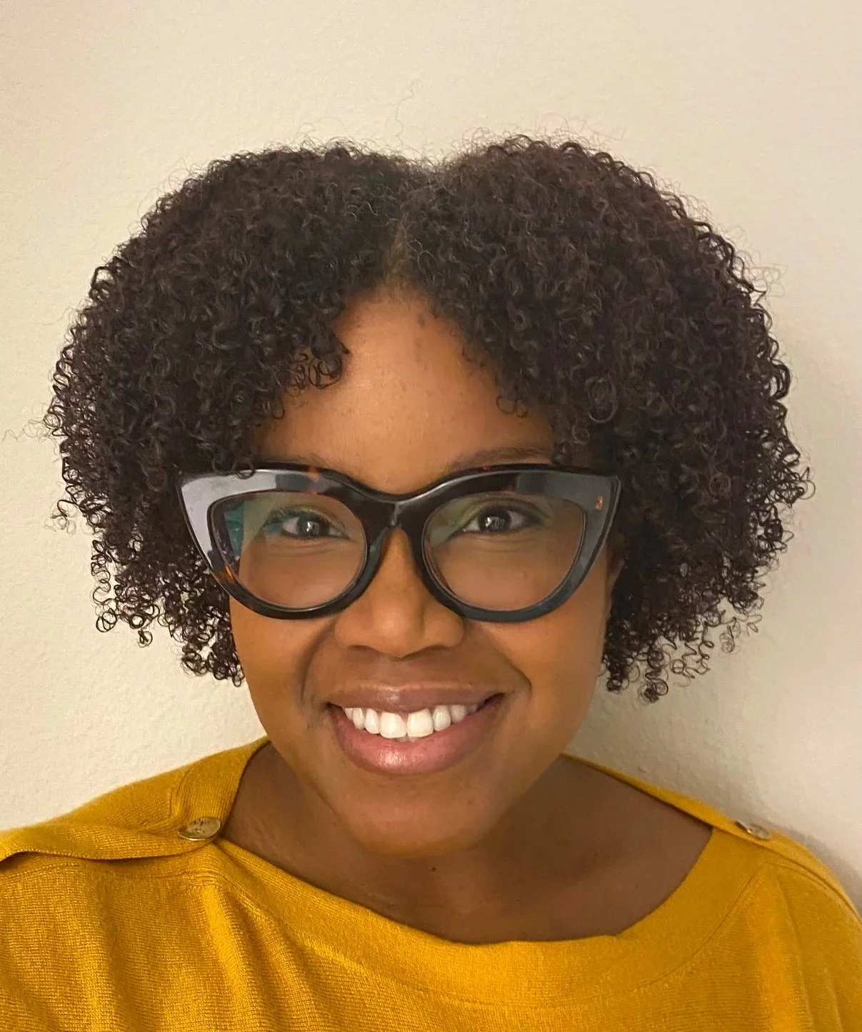 A woman with short curly hair, wearing large tortoiseshell glasses and a yellow top, smiling at the camera against a plain background.