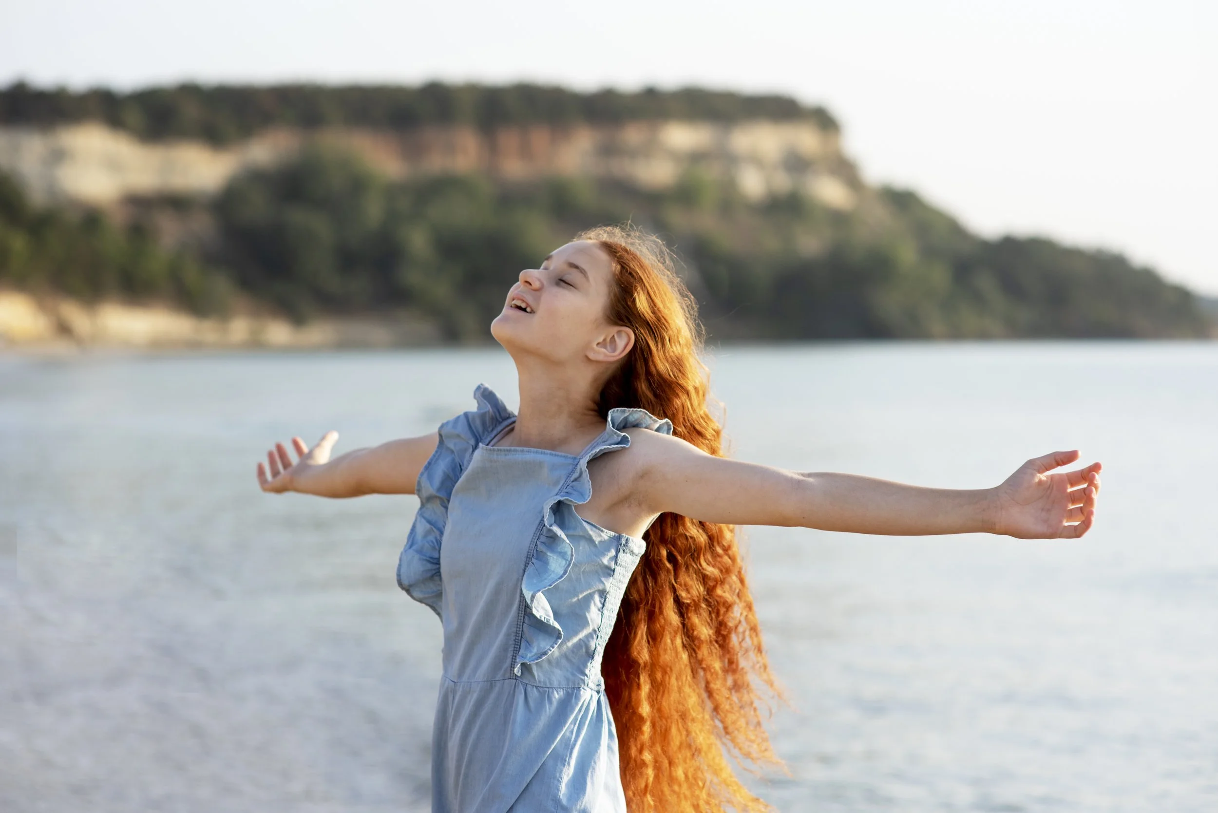A woman with long red hair smiling with her arms outstretched near a body of water, with cliffs and trees in the background.