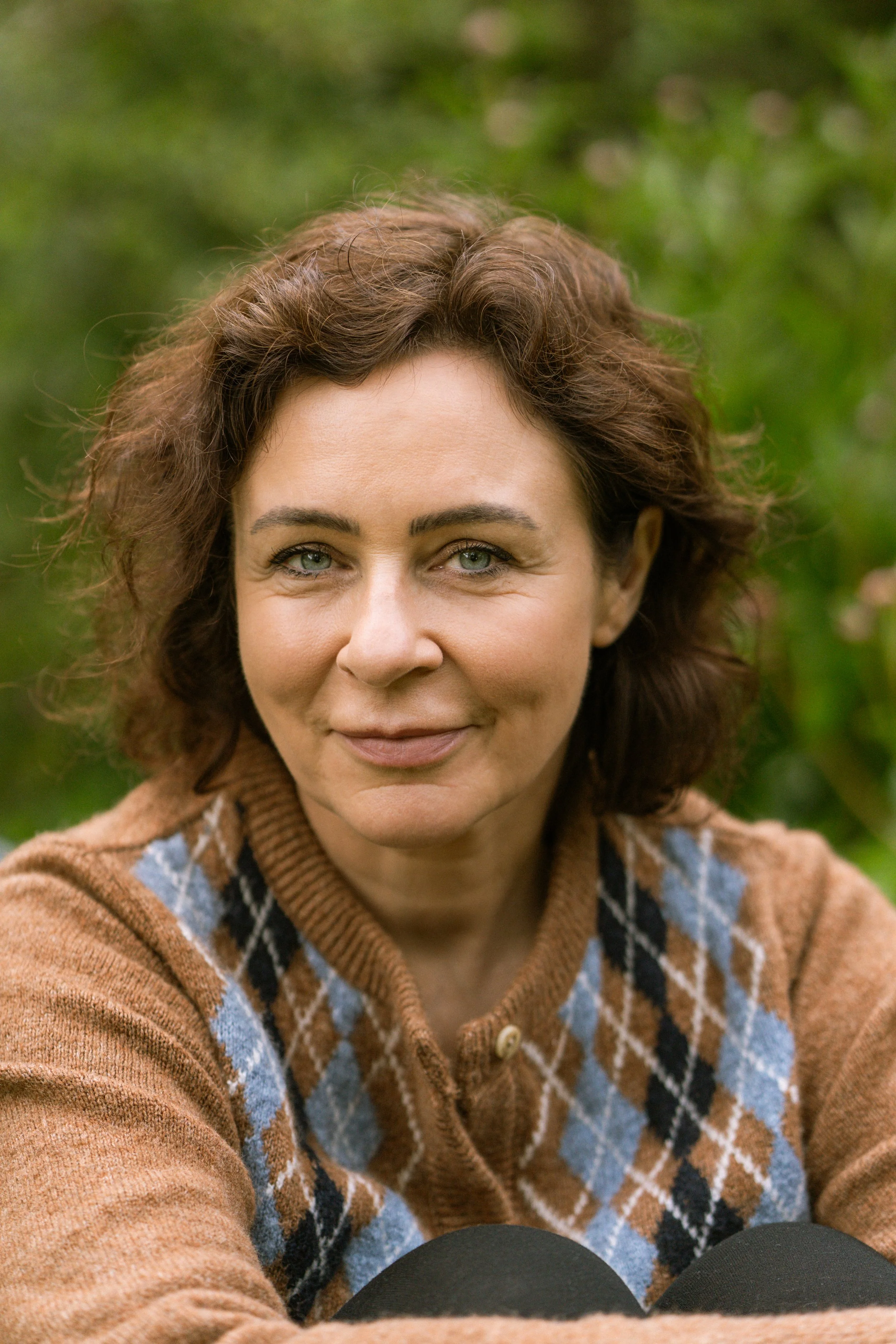 Close-up of a middle-aged woman with curly brown hair and blue eyes, smiling outdoors with a blurred green background, wearing a brown argyle sweater.
