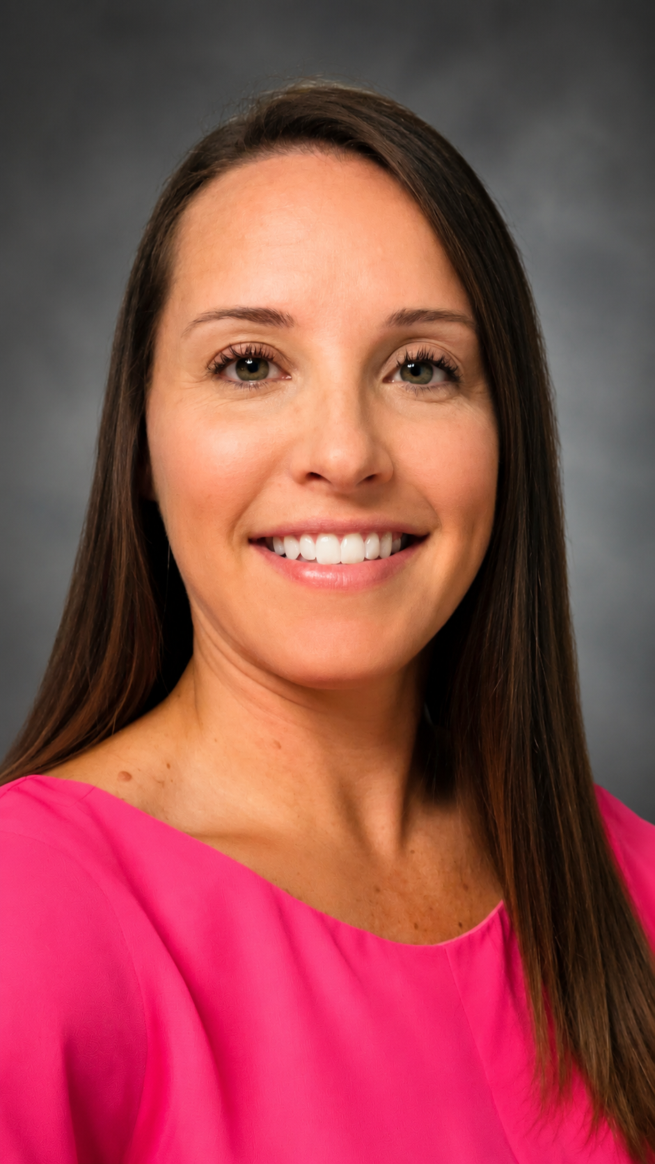 A woman with long brown hair wearing a pink top smiling at the camera against a gray background.