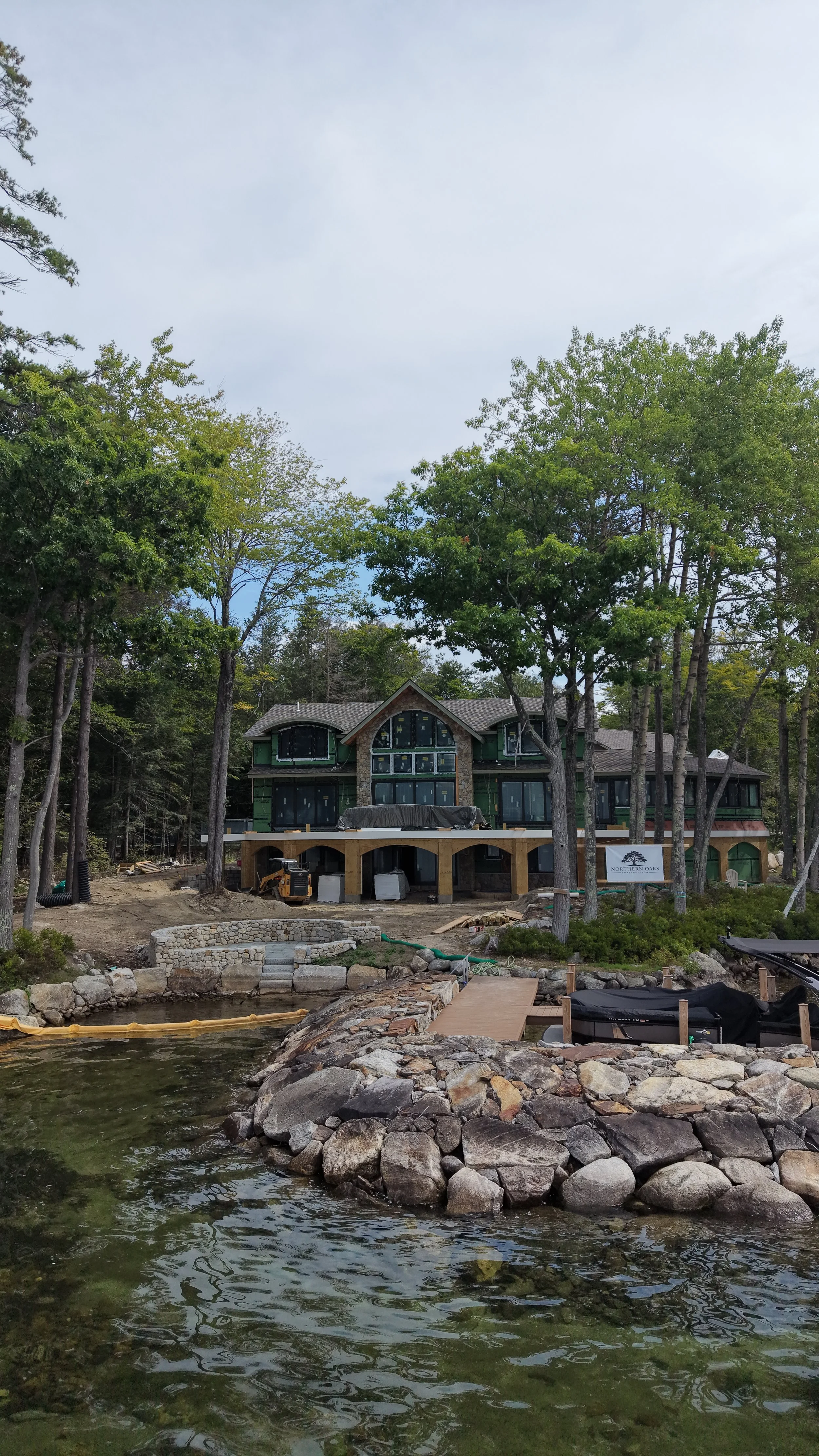 A house under construction near a body of water, surrounded by trees, with construction equipment and materials visible.