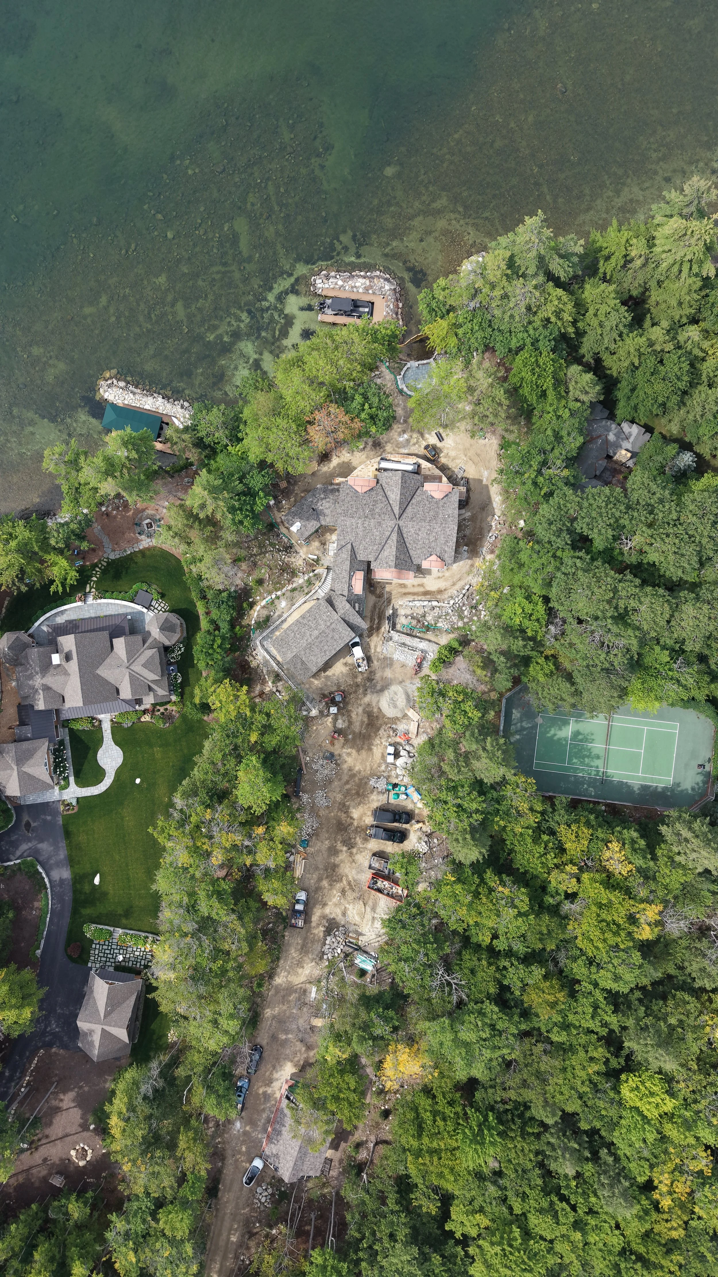 Aerial view of a residential backyard under construction, showing a large house, surrounding trees, a tennis court, a dirt driveway with parked cars, and a swimming pool surrounded by trees.