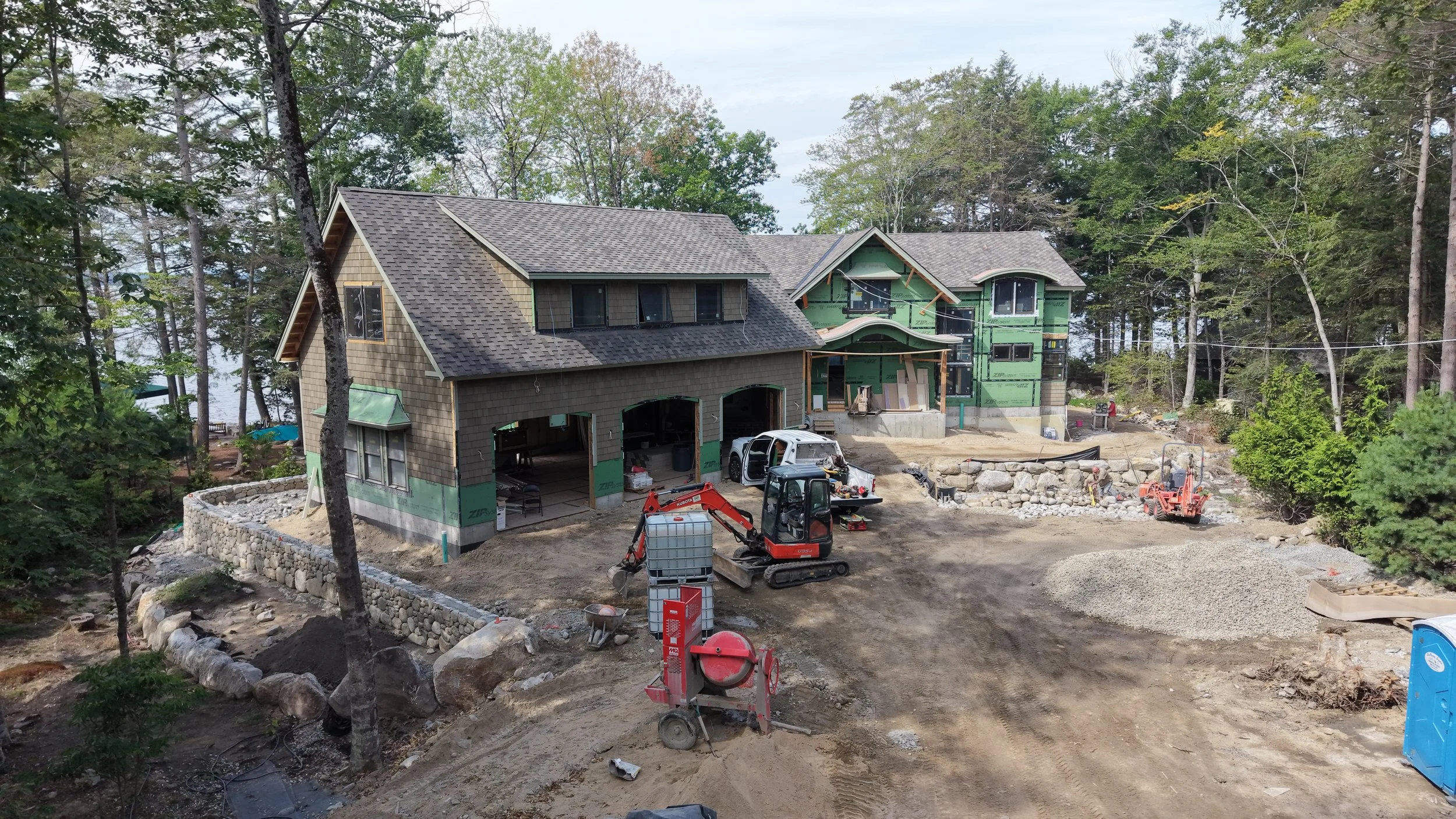A house under construction surrounded by trees with a visible garage and multiple construction tools and equipment, including a small excavator, a work truck, and a portable toilet.