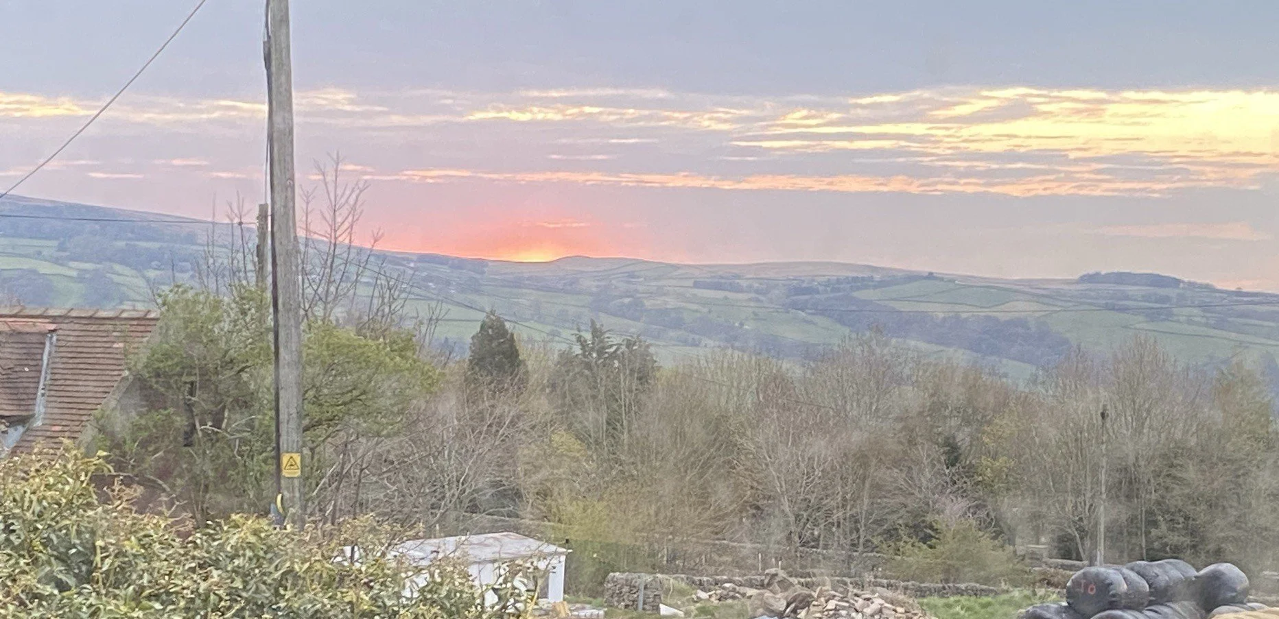 Sunset over rolling hills with pastel-colored sky, bare and green trees, and rooftops in a rural landscape.