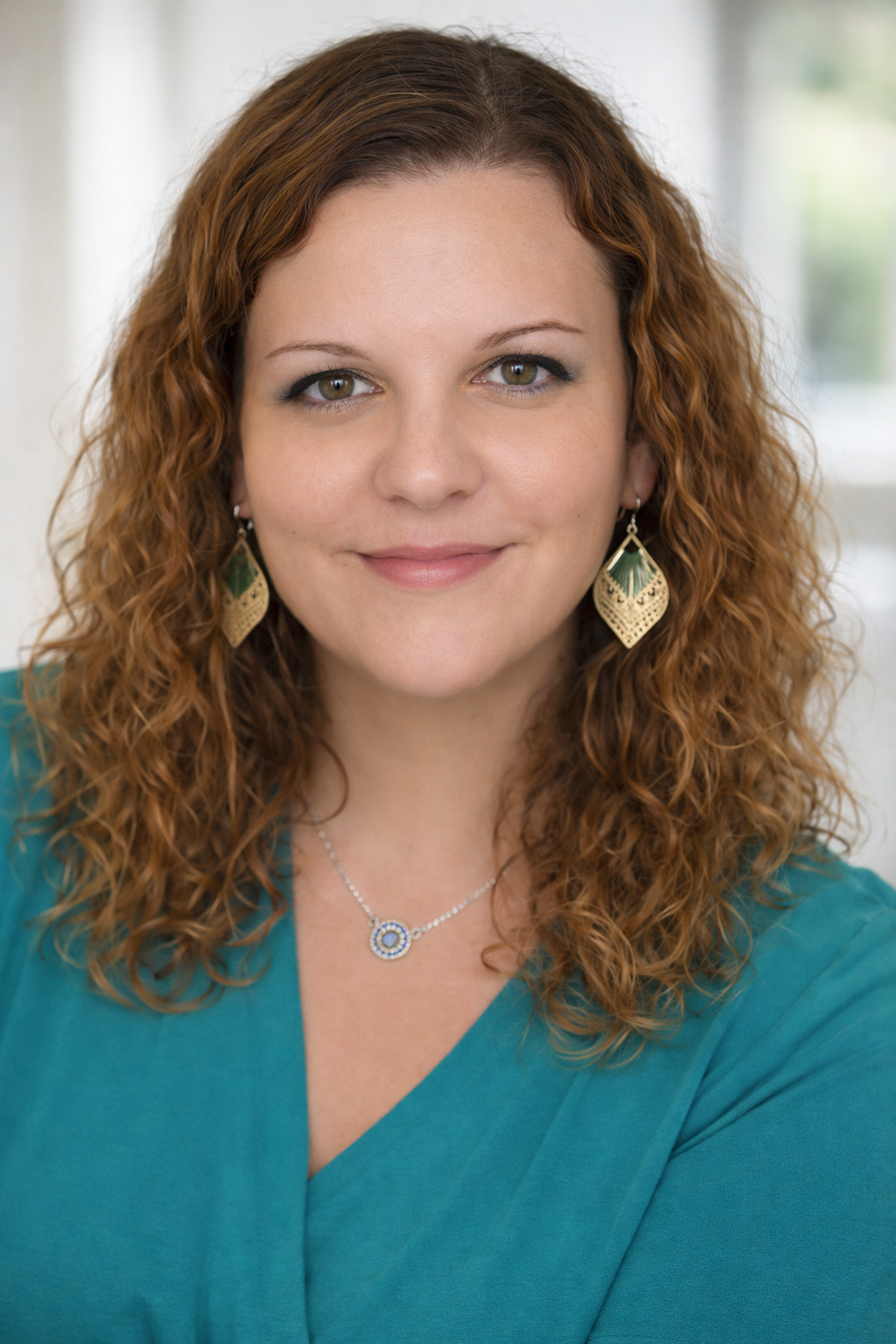 Jeune femme aux cheveux bouclés roux clair, portant des boucles d'oreilles dorées avec des détails verts, une chaîne avec un pendentif bleu et un haut turquoise, souriant doucement sur un fond lumineux