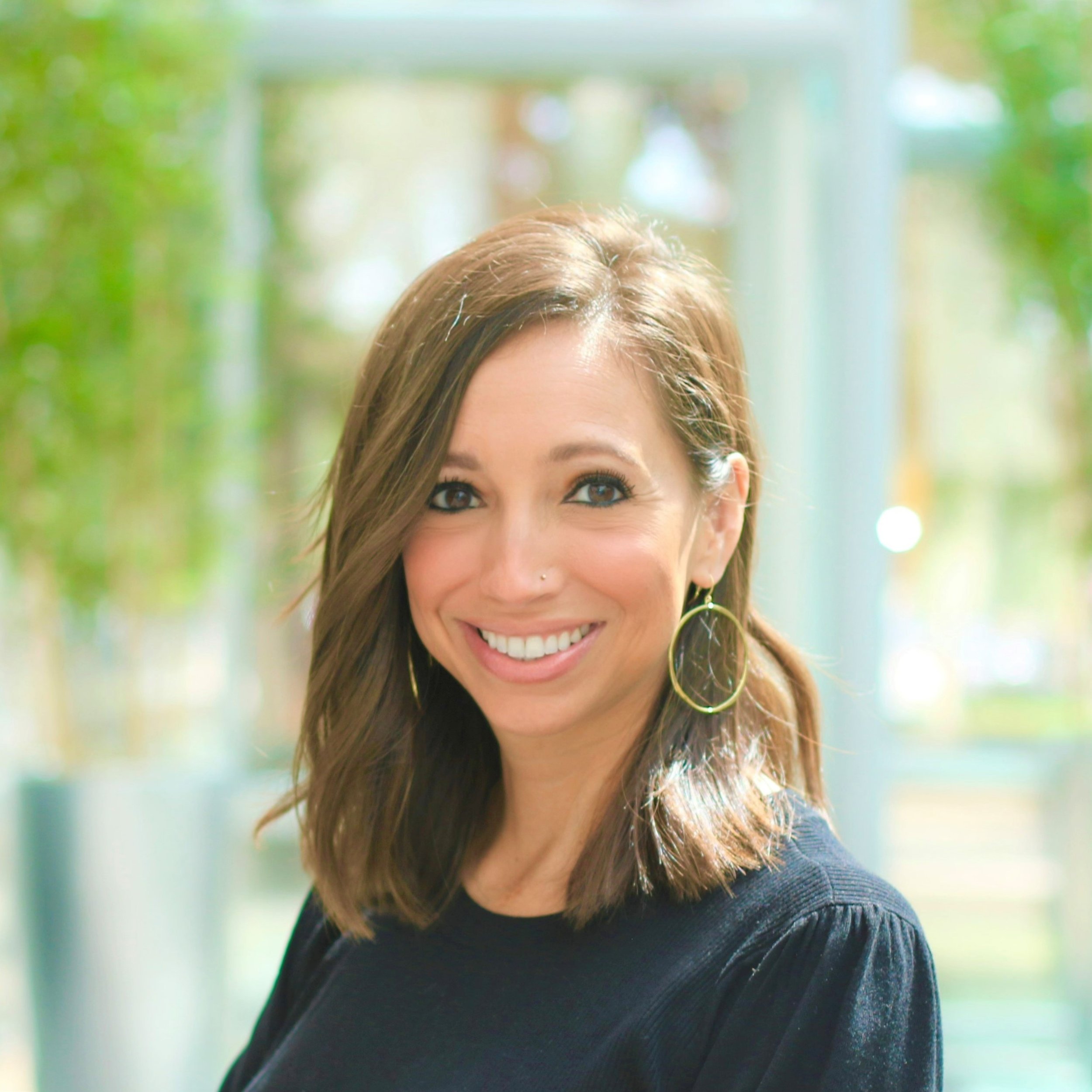 Close-up of a smiling woman with brown hair, wearing makeup, ear jewelry, and a black top, outdoors with blurred trees and natural light in the background.