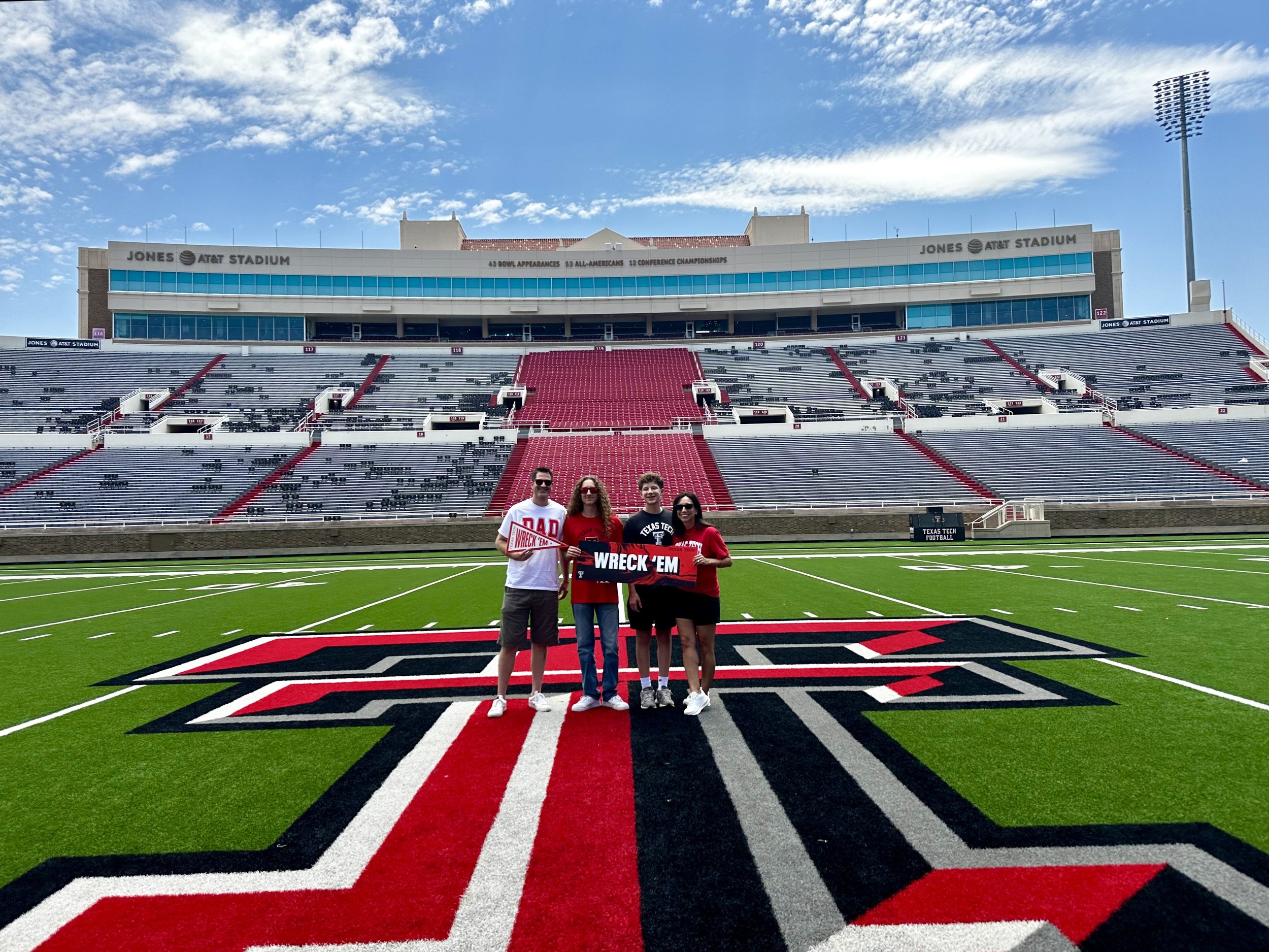 Four people standing on the field of a large football stadium, in front of the large logo on the field, with the stadium seats and a blue sky with clouds in the background.
