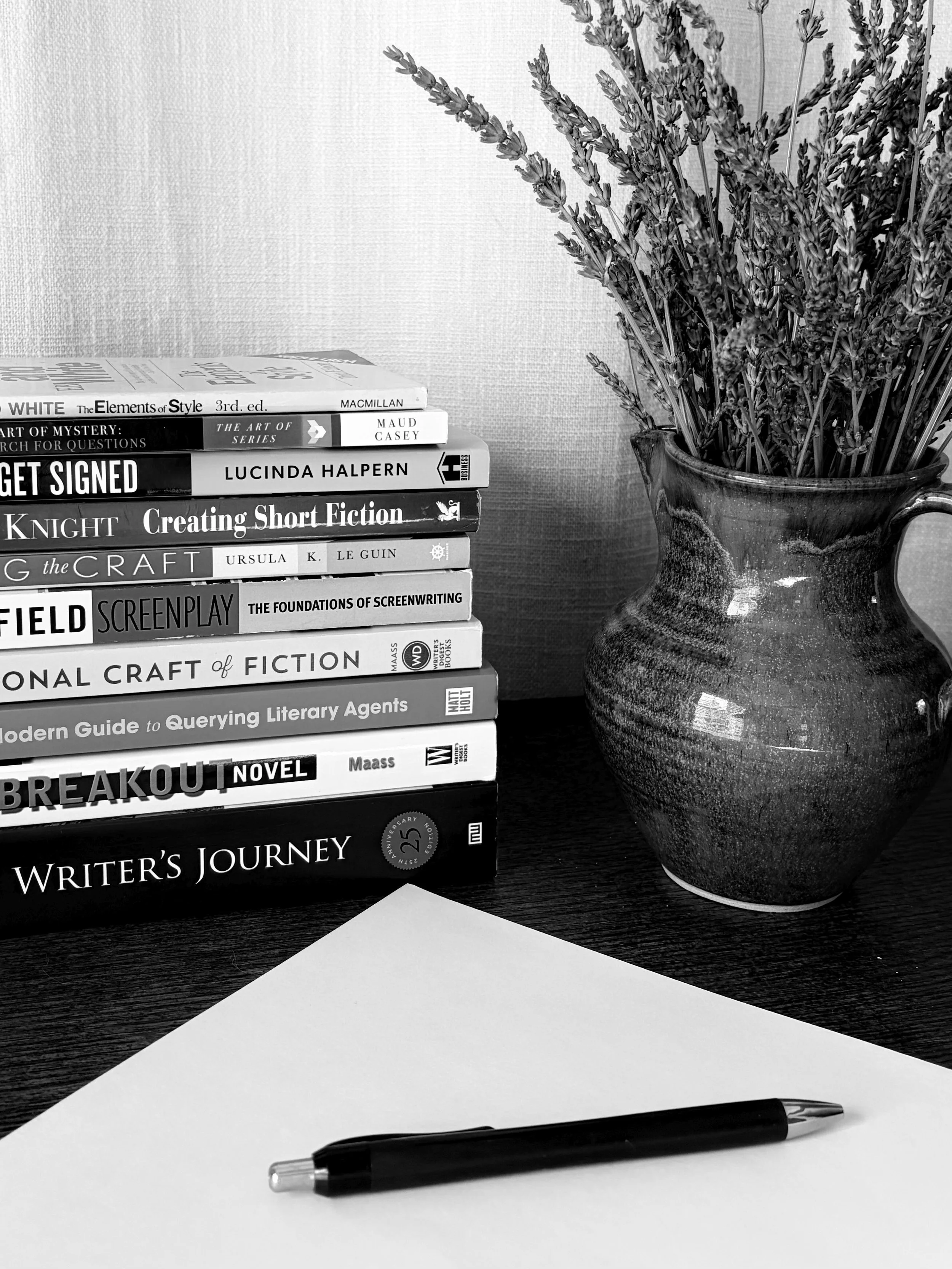 A black pen resting on a white sheet of paper on a dark surface. Behind the paper is a stack of books about writing and storytelling, and a large ceramic vase with dried lavender flowers.