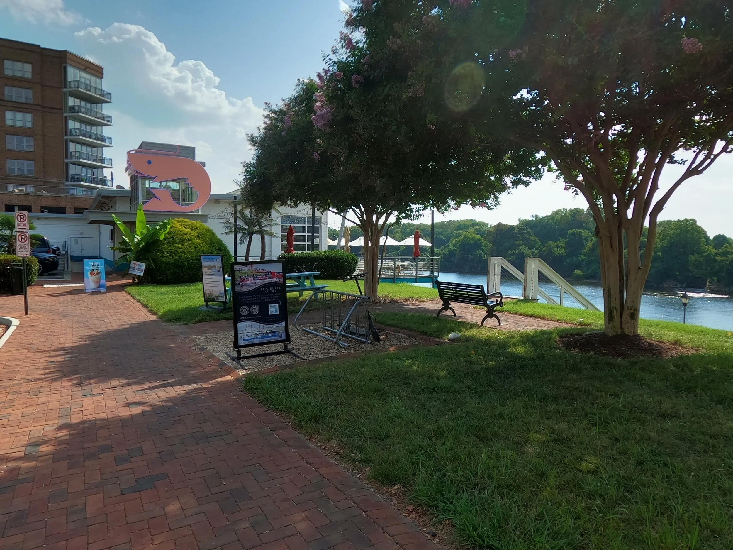 Outdoor scene with brick pathway, green grass, trees, umbrellas, benches, buildings, and a body of water in the background.