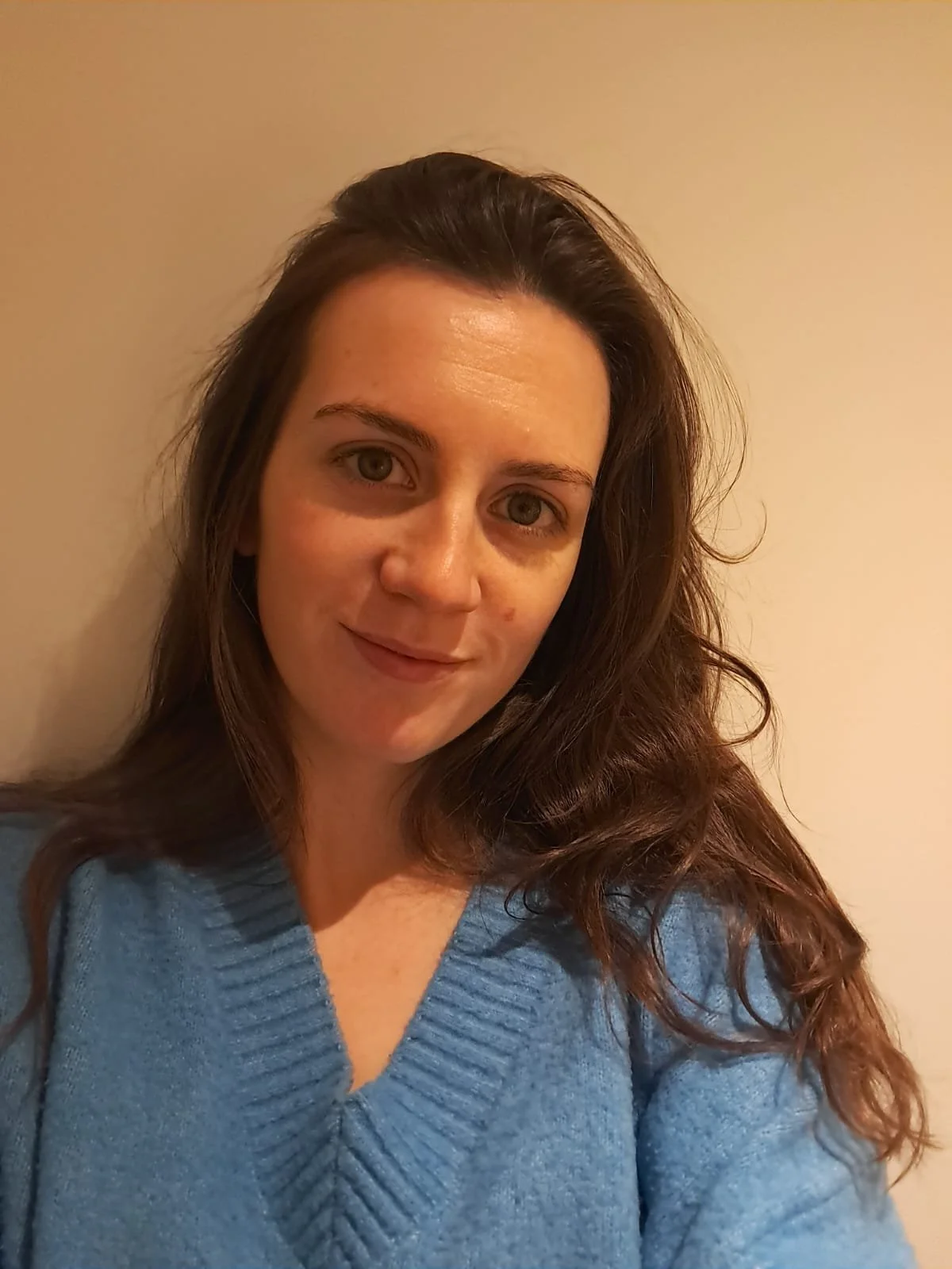 A woman with long brown hair wearing a blue sweater, standing against a plain light-colored wall, smiling slightly.