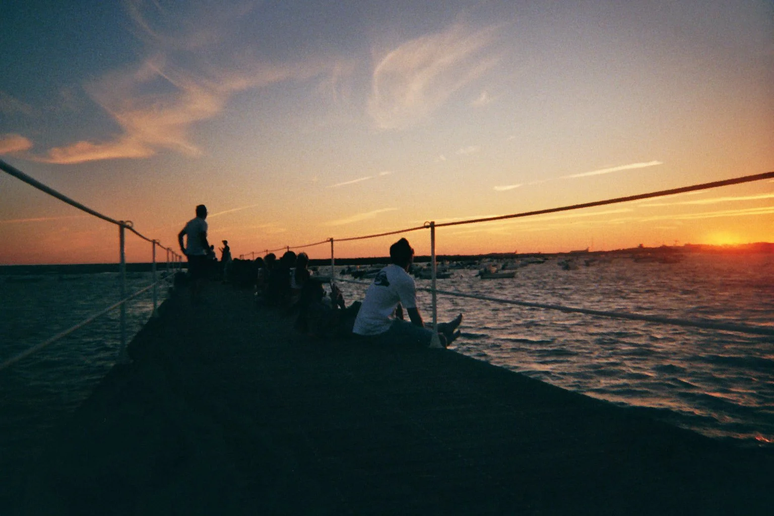 Folk sitter på en brygge under solnedgang, langs kysten, med folk og seilbåter i bakgrunnen.