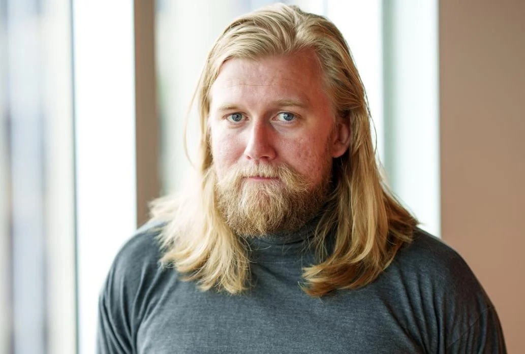 Et portrett av en mann med langt blondt hår og skjegg, iført en grå genser, som står innendørs foran et vindu.