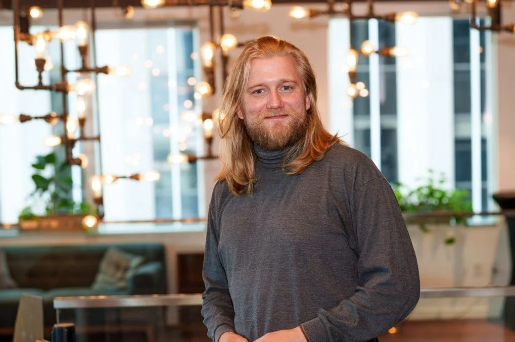 En mann med langt, blond hår og skjegg, iført grå genser, smiler innendørs i et moderne, lyst rom med store vinduer og dekorative lamper.