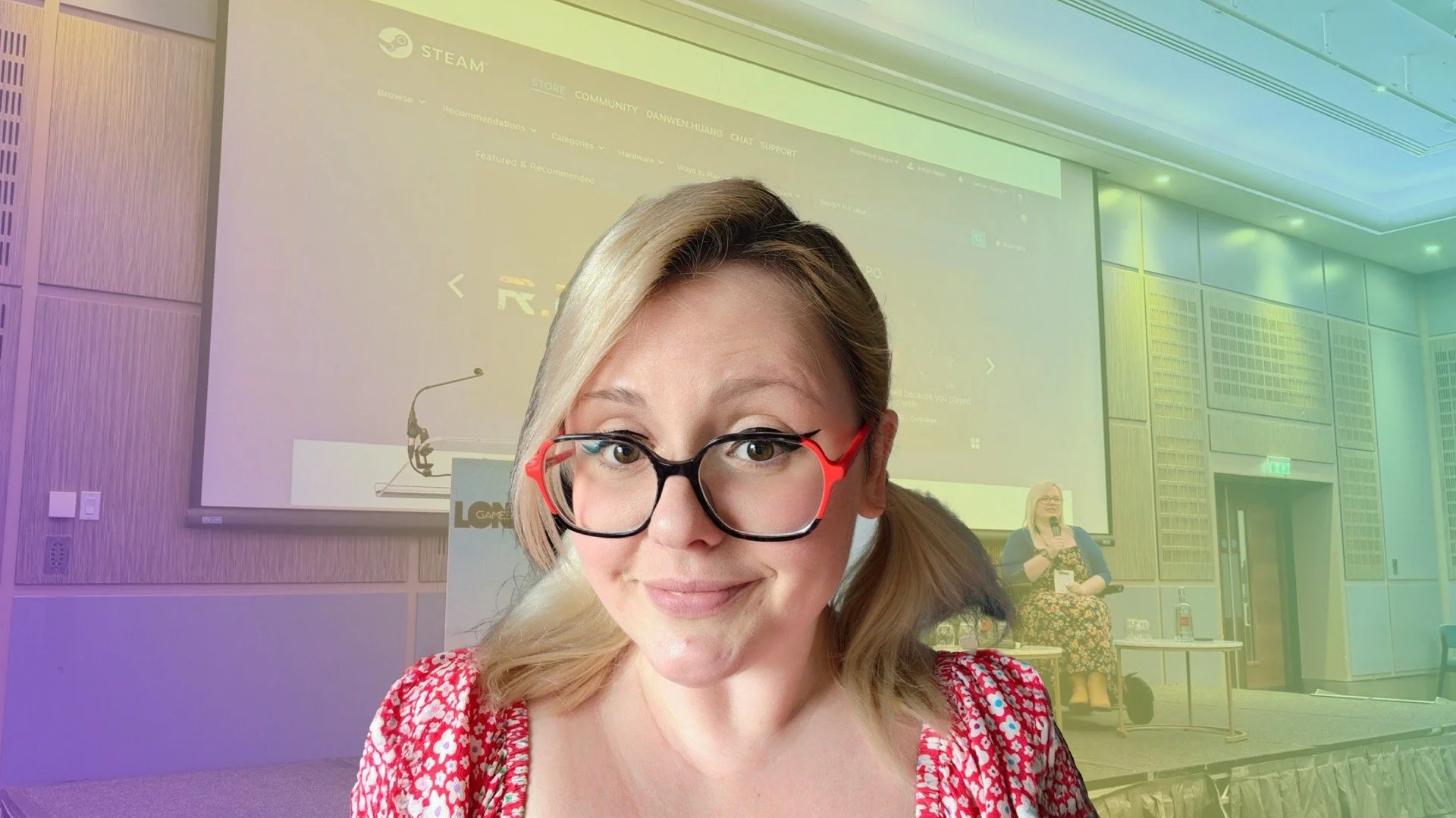 Close-up of a woman with glasses smiling in front of a conference room screen, with a woman in the background speaking into a microphone.