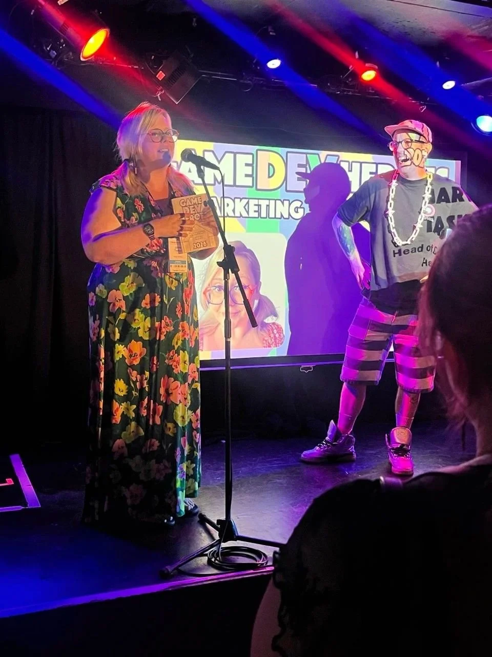 A woman in a floral dress holding a certificate and speaking at a microphone on stage with two people dressed in costumes, during an event called Game Developers Conference 2016.