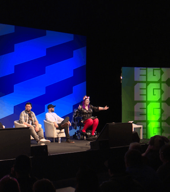 A panel of four people on stage during a conference or discussion. One woman is speaking or presenting, seated in a wheelchair. The other three men are sitting in chairs listening. The background features colorful geometric designs and a blue and green patterned wall.