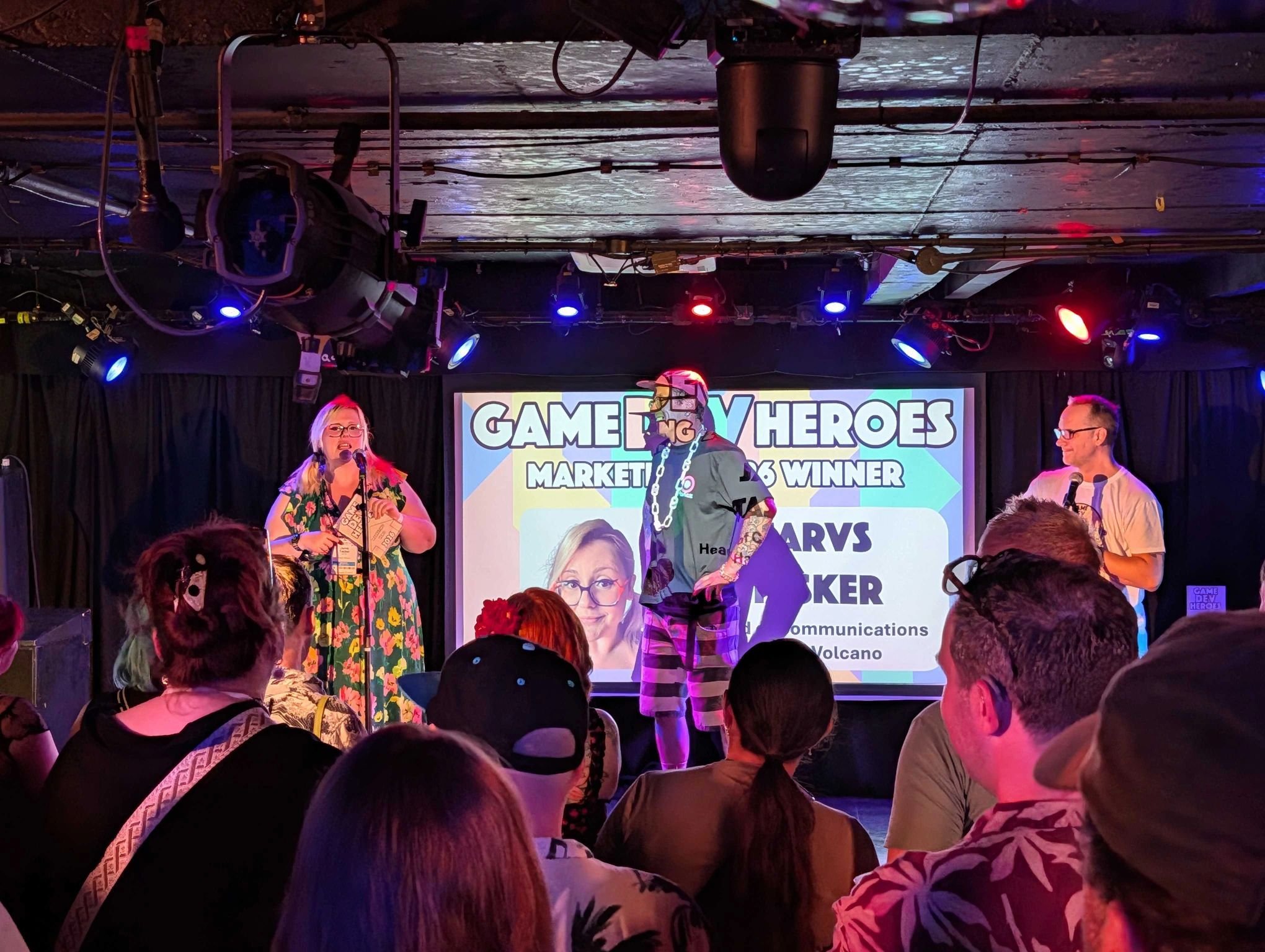 People standing and sitting in an audience watching a stage presentation with a large screen behind them. The stage features a woman speaking into a microphone, a person dressed in a costume with a chain, and a man with glasses. Four stage lights hang from the ceiling.