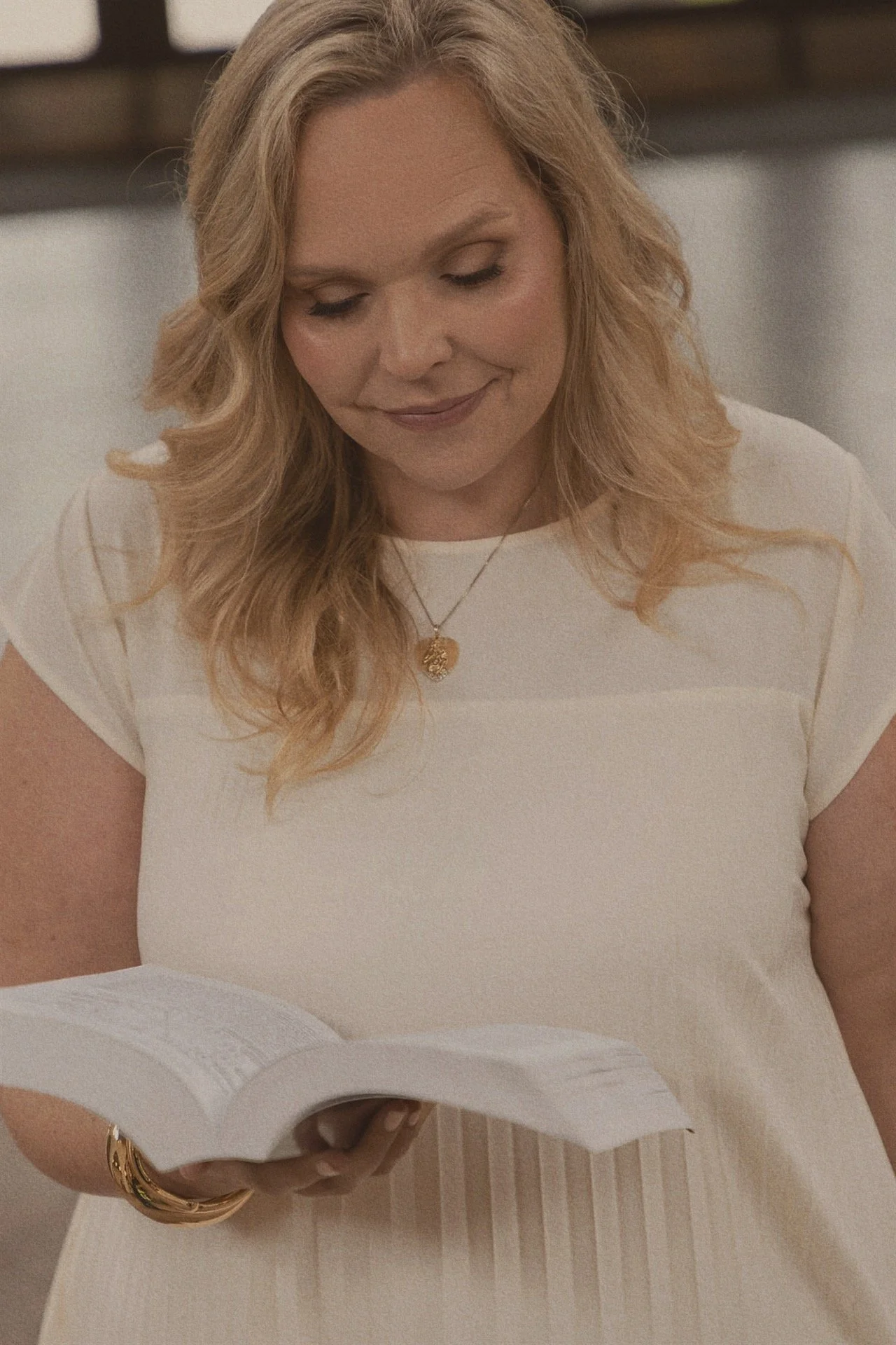 A woman with wavy blonde hair, wearing a white top and gold jewelry, is smiling while looking down at a piece of paper in her hand.