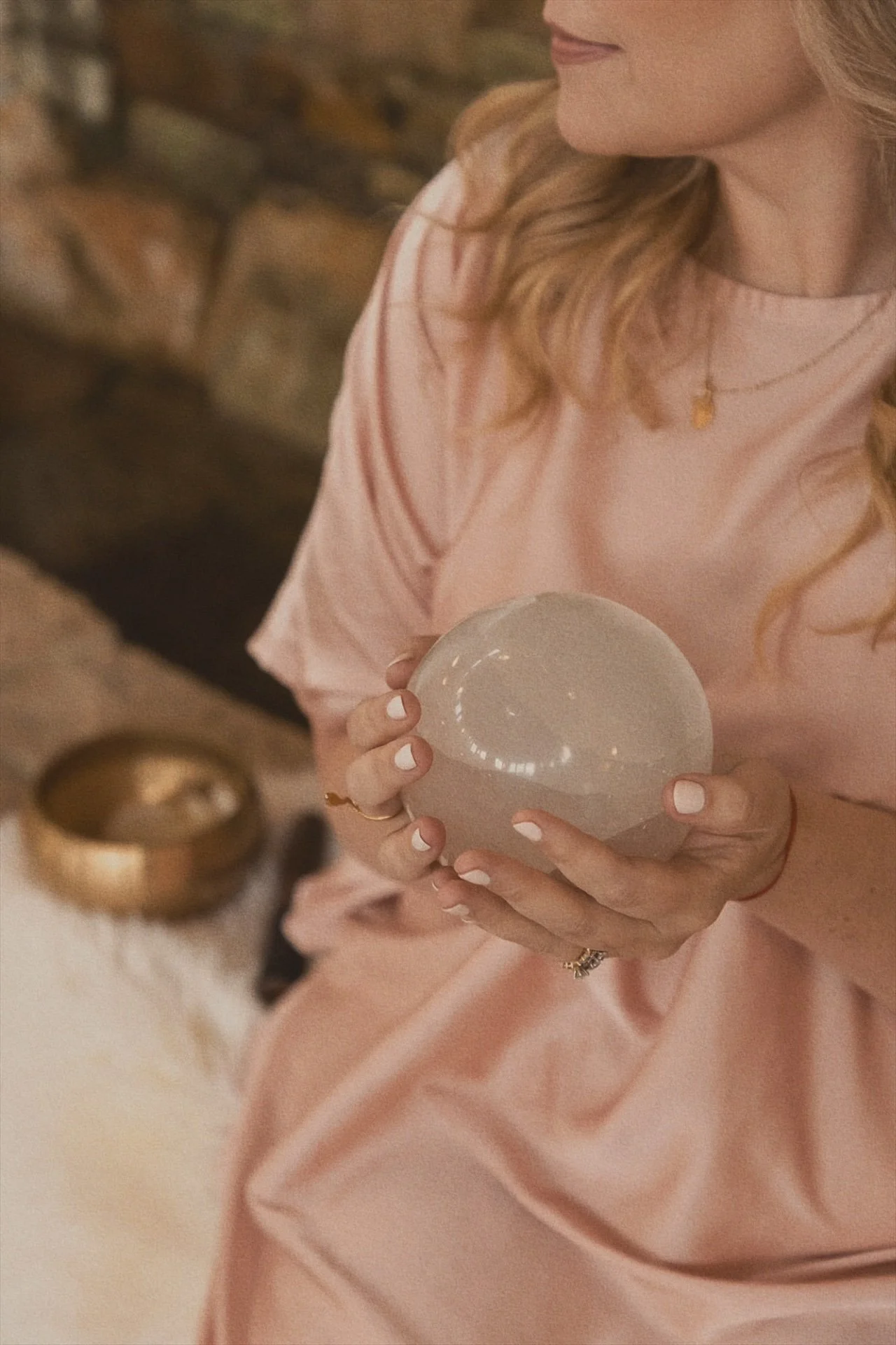 A woman in a pink satin blouse holding a white bowl with her left hand, with layered blonde hair, a gold necklace, and a ring on her finger.