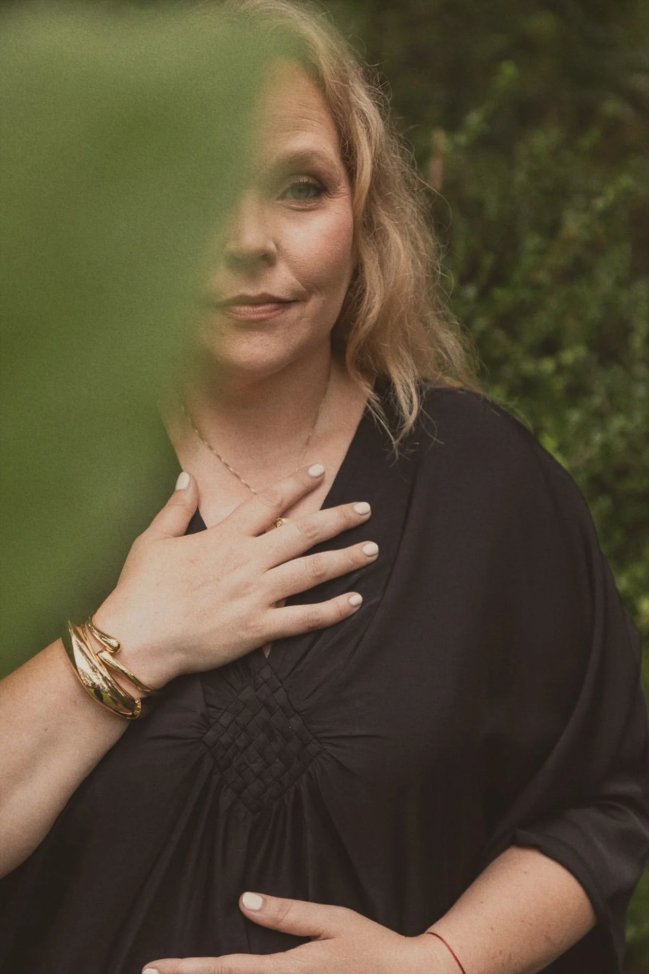 A woman with curly blonde hair, wearing a black dress, gold bracelets, a ring, and a necklace, is touching her chest with one hand and resting her other hand on her abdomen. She is partially obscured by a green leaf in the foreground, set against a blurred background of greenery.