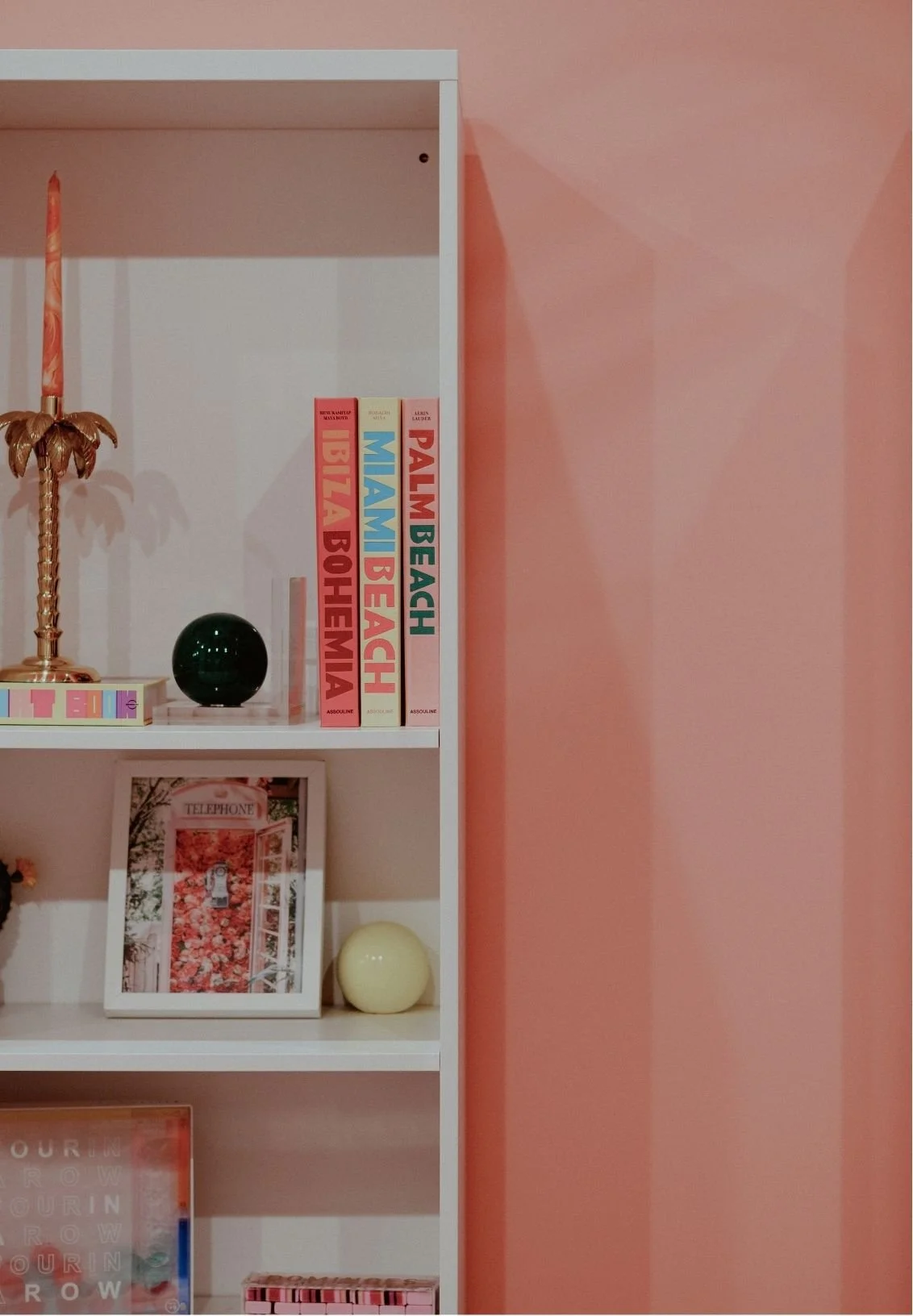 Pink built-in bookshelf styled with colorful coffee-table books, a palm tree candle, decorative spheres, and framed floral artwork.