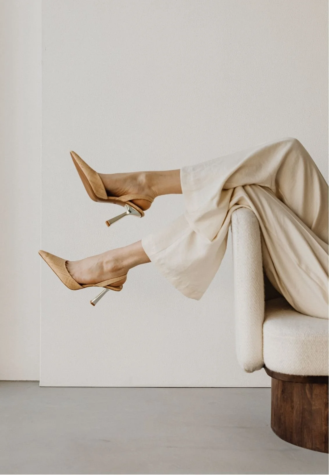 Woman in flowing cream pants and pointed beige high heels reclining on a white armchair.