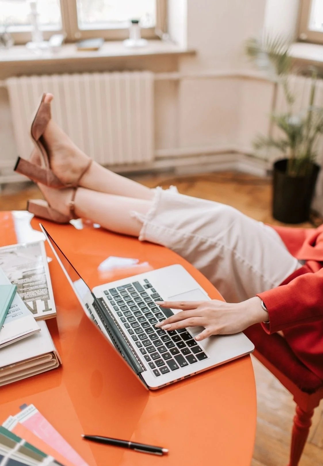 Woman working on a laptop at an orange table with papers and a pen, wearing a red sweater and heels