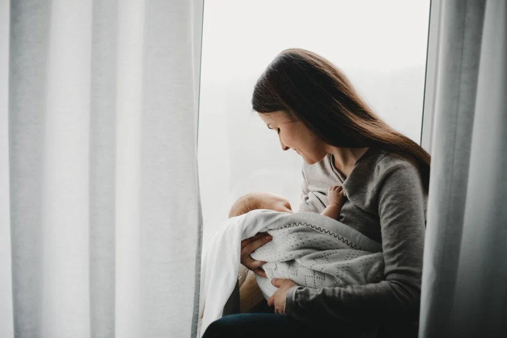 Mother breastfeeding her baby while sitting by a bright window