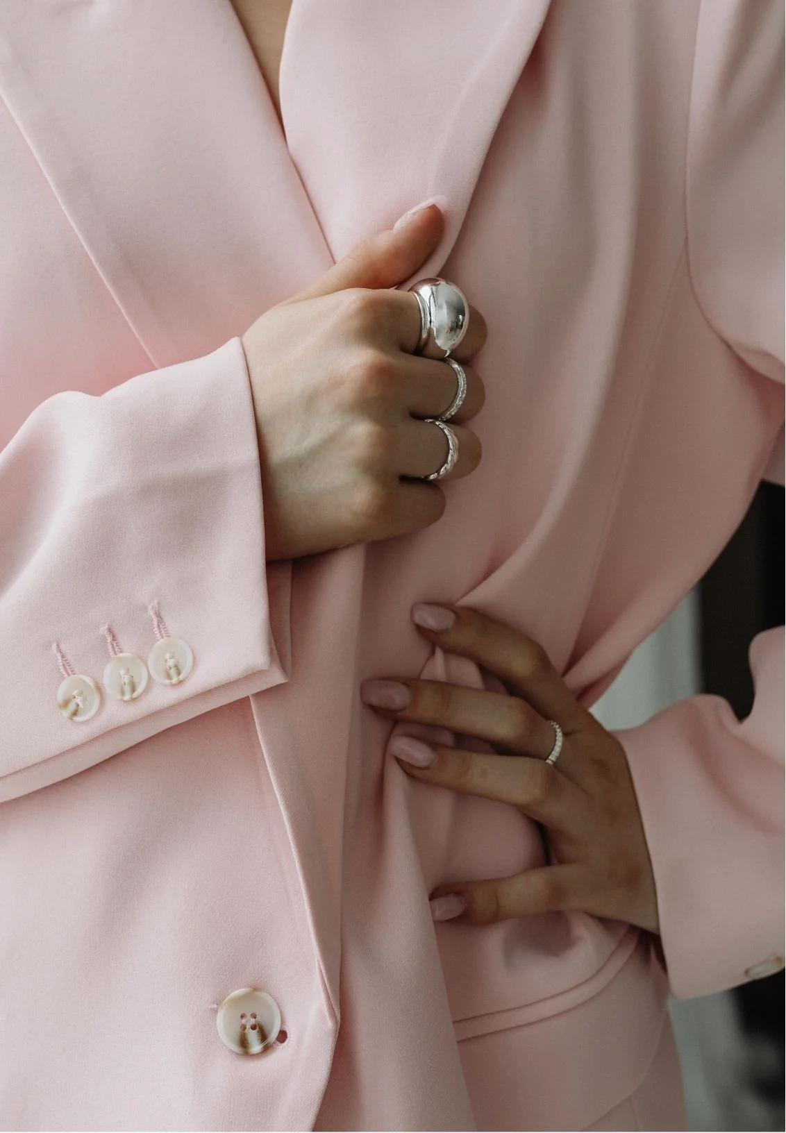 Close-up of a person wearing a blush pink suit jacket with white buttons, flowing lapels, and silver rings.