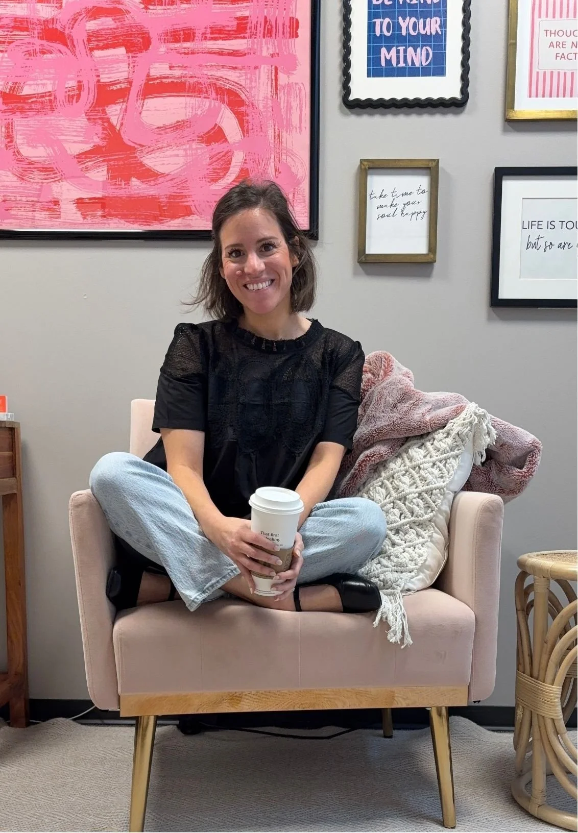 Jen Greenberg, a therapist based in North Dallas / Plano, smiling and sitting cross-legged in a pink armchair, holding a coffee cup beside framed motivational artwork.